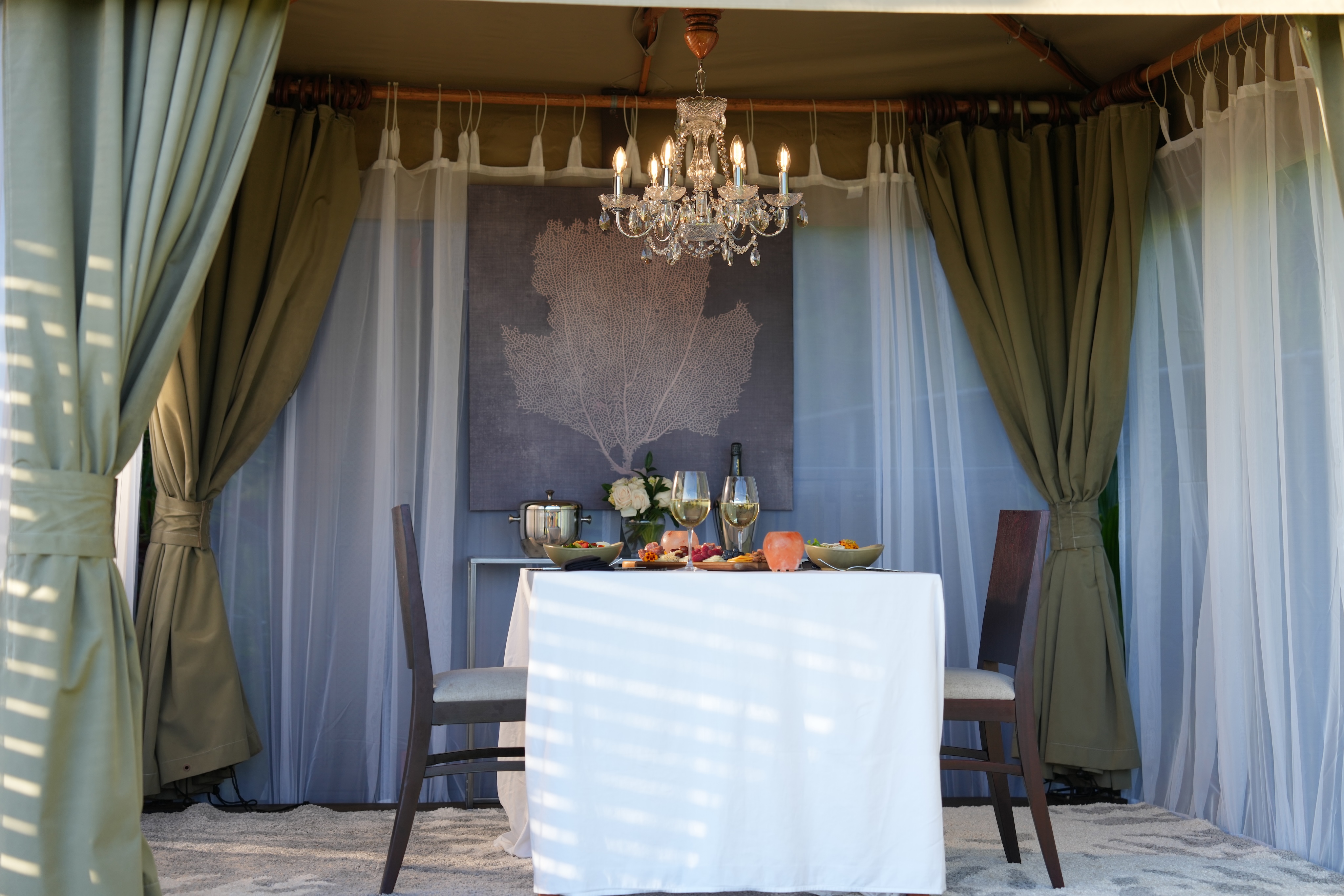 A small dining table set for two under a chandelier, surrounded by green curtains and a large wall decoration, with wine glasses, plates of food, and a bottle displayed. The setting exudes an oceanfront ambiance reminiscent of cozy cabanas.