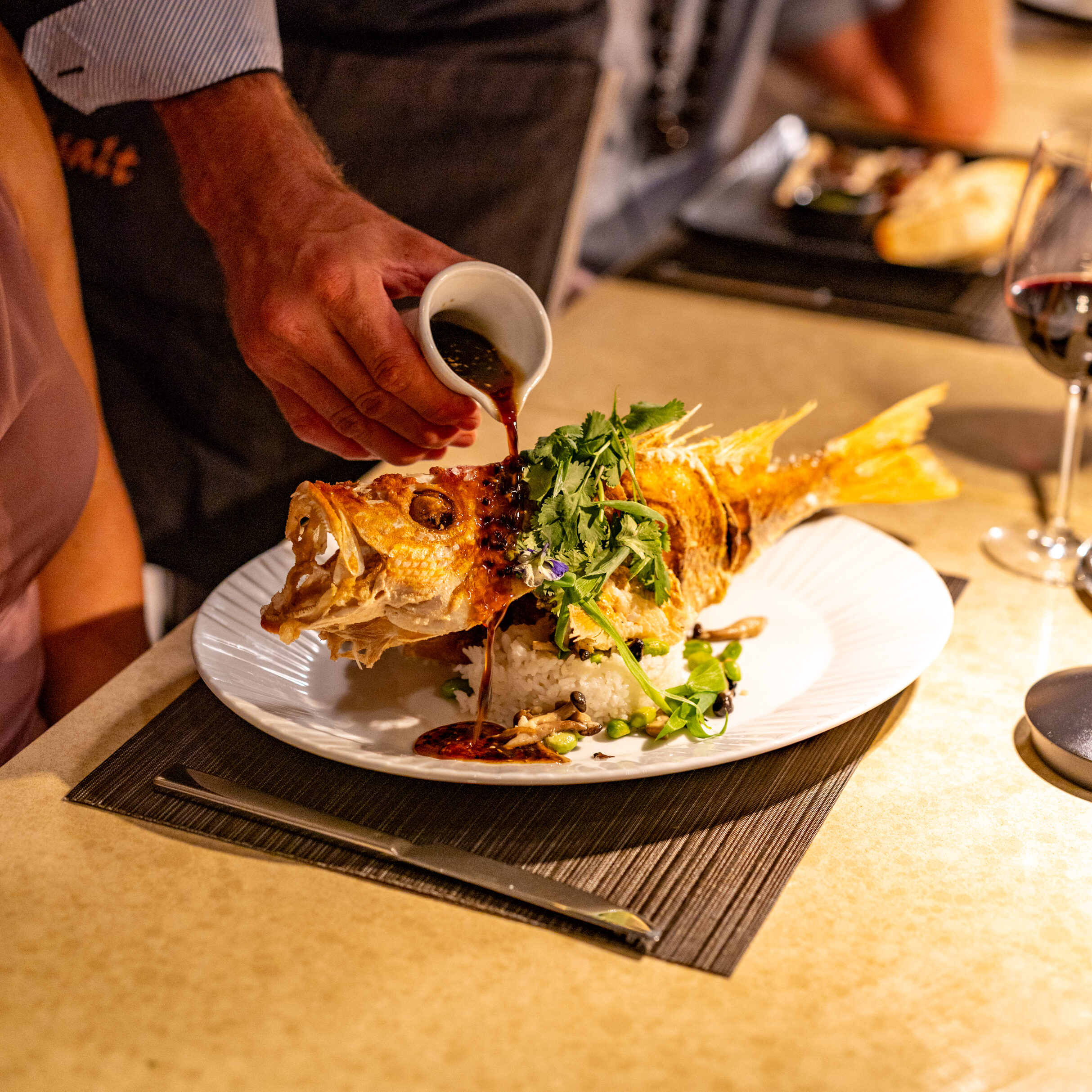 A server pours sauce over a whole cooked fish garnished with herbs on a white plate in a restaurant setting.