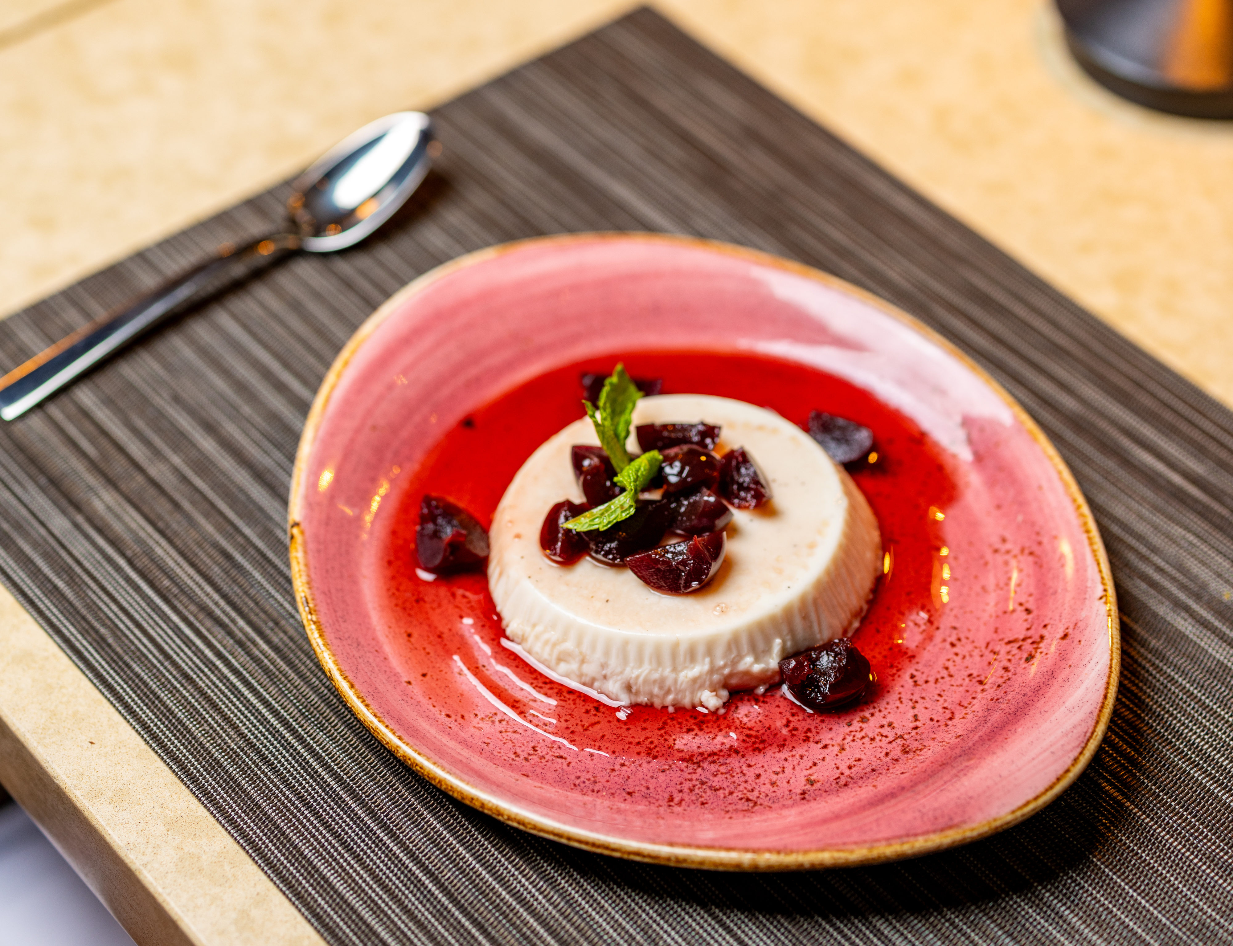 A plate of panna cotta topped with cherries and mint sits on a pink dish next to a spoon on a dark placemat, making it a standout choice for any dessert menu.