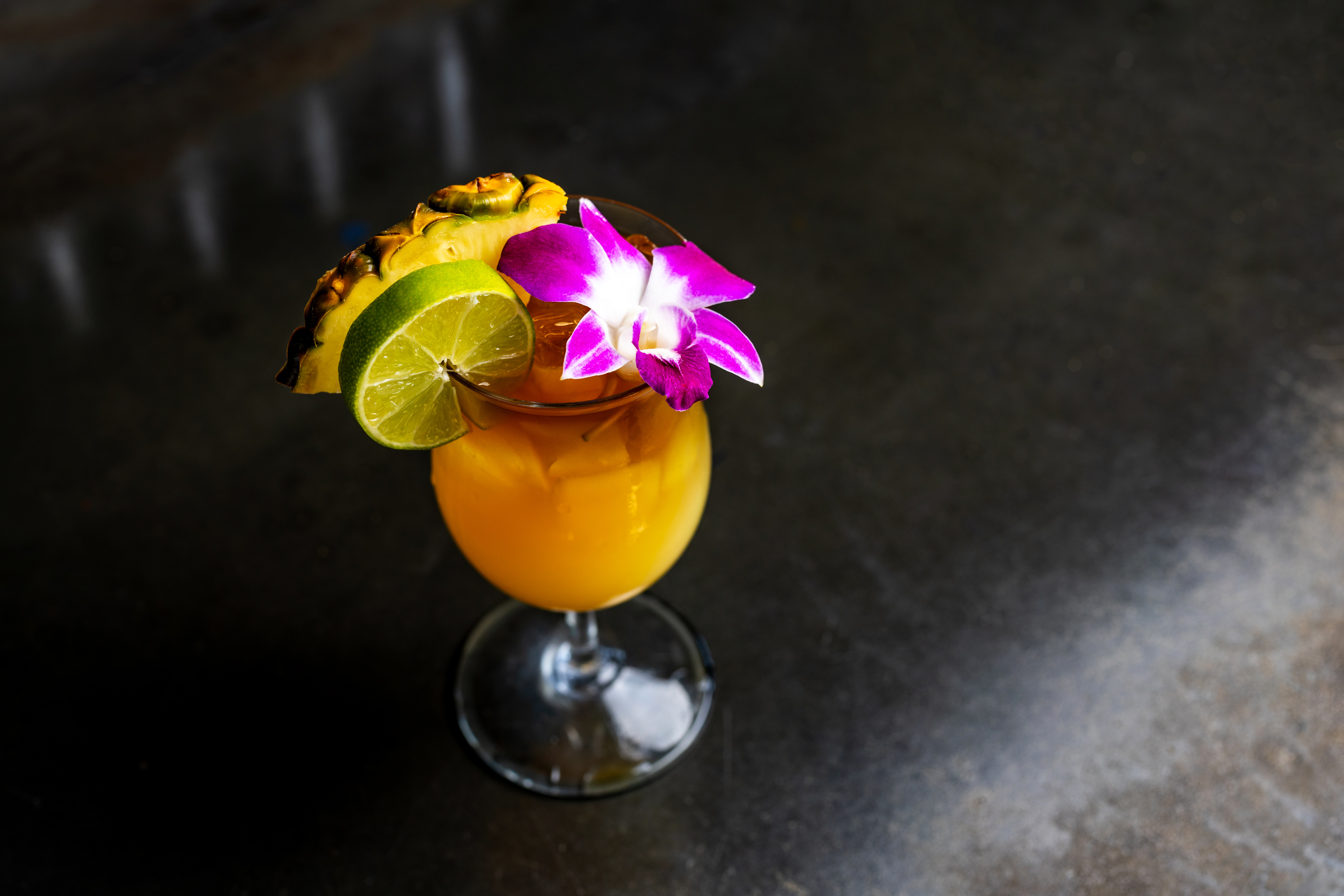 A tropical cocktail from our beverage menu, garnished with a lime slice, pineapple, and a purple orchid flower, served on a dark surface.
