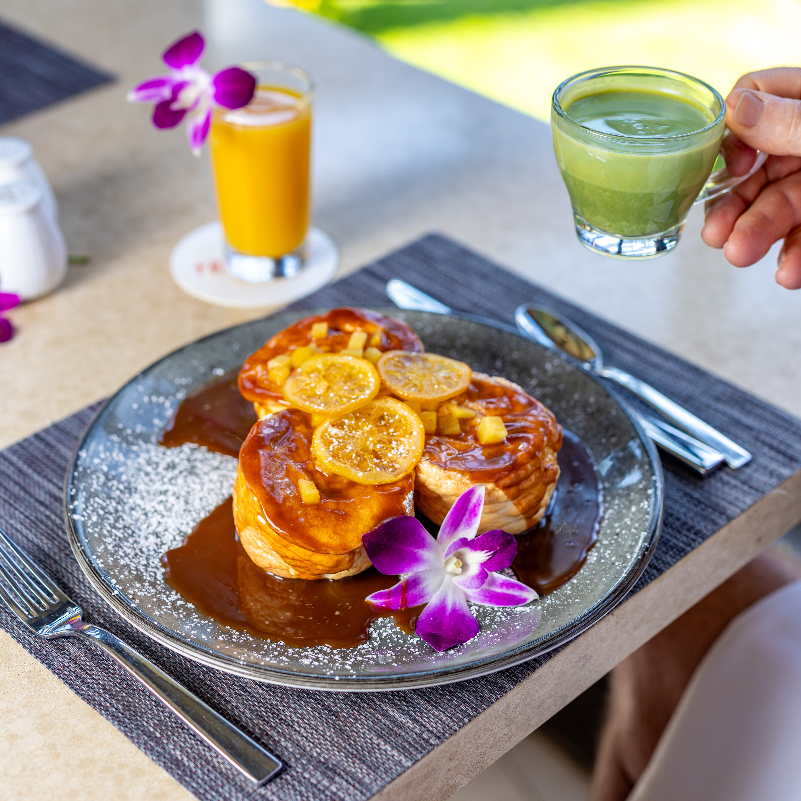 A plate of pancakes topped with caramel sauce and lemon slices, garnished with purple orchids, sits beside a glass of orange juice and a hand holding a cup of green beverage—an inviting Breakfast Menu delight.