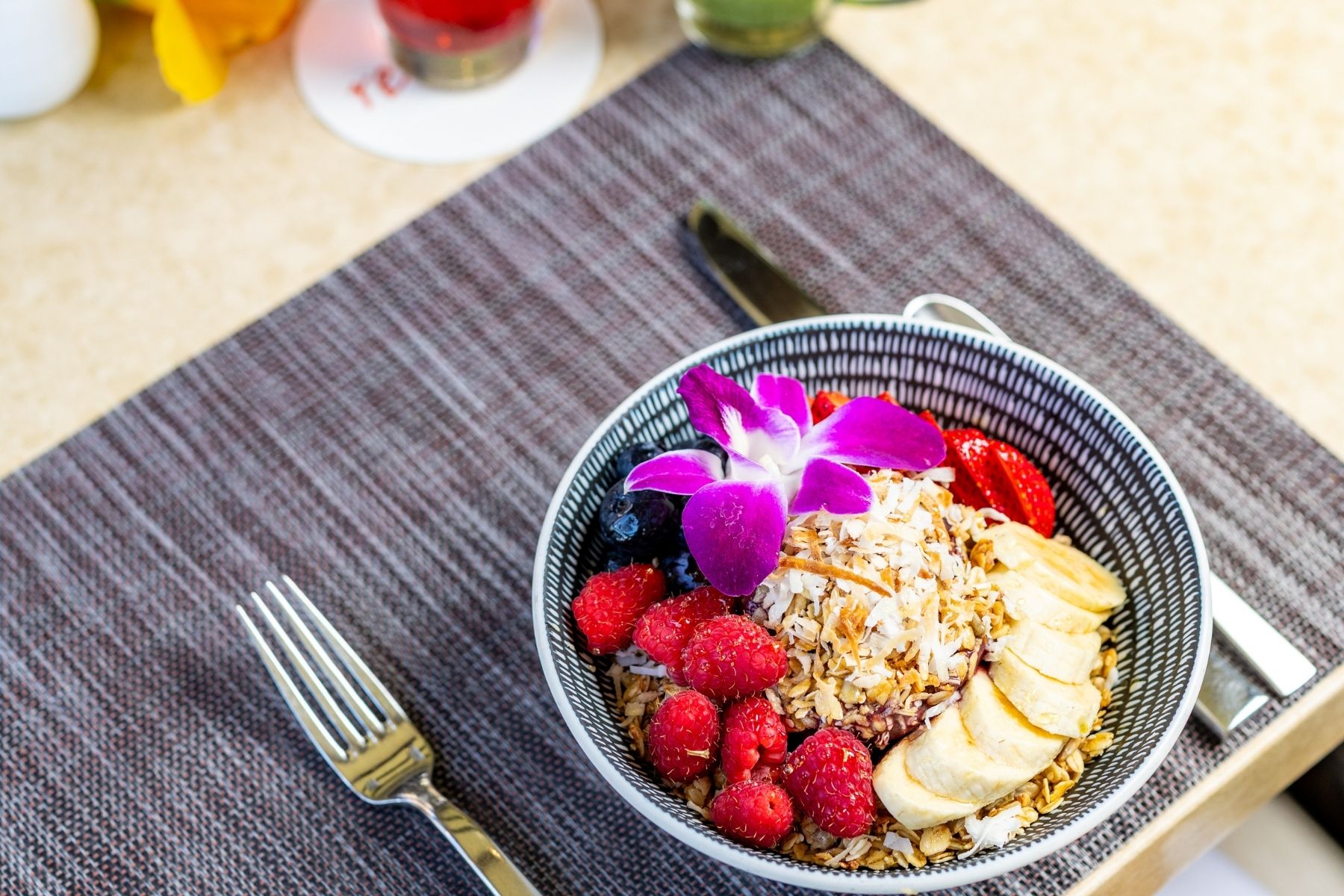 A bowl of granola topped with sliced banana, raspberries, strawberries, coconut, and a purple orchid flower is presented on a placemat with a fork and knife beside it—a vibrant addition to any breakfast menu.