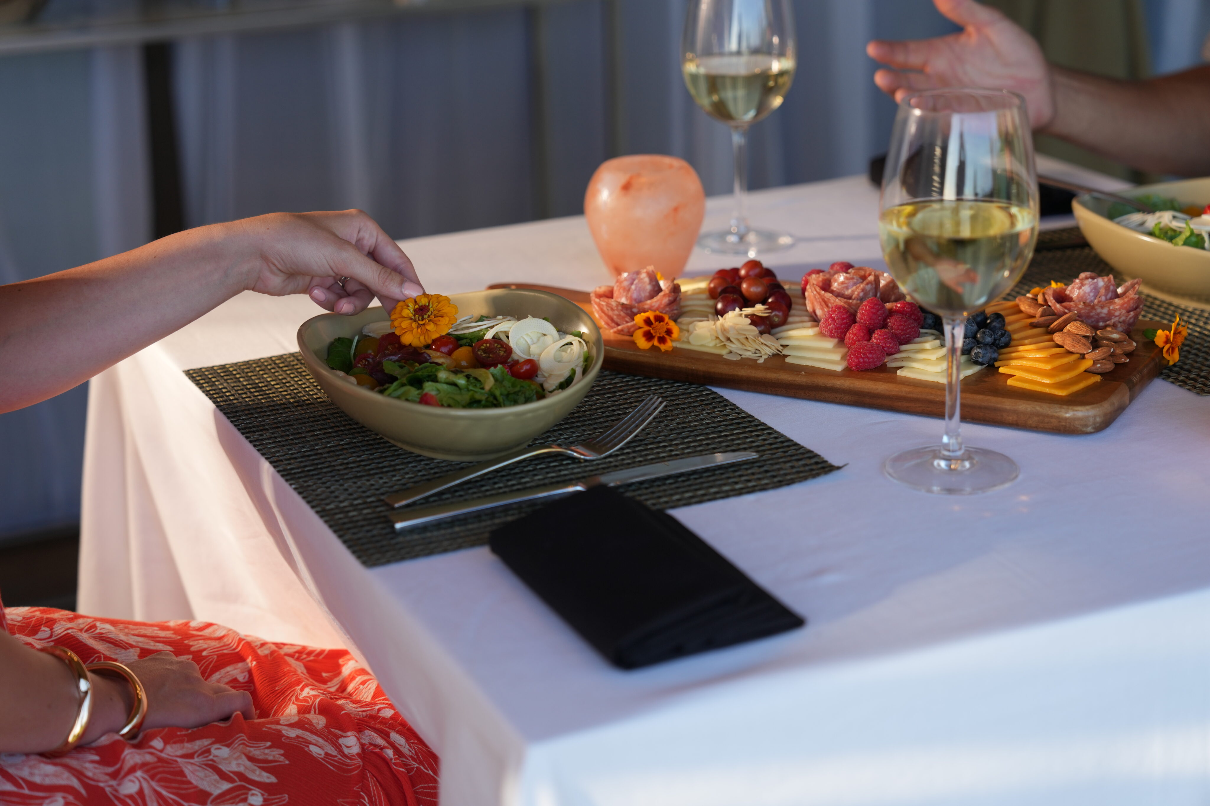 Two people dining. One hand reaches for salad, while the other points to a charcuterie board with fruit, cheese, and meat on a white table with wine glasses—truly a picture-perfect page from an elegant home gathering.