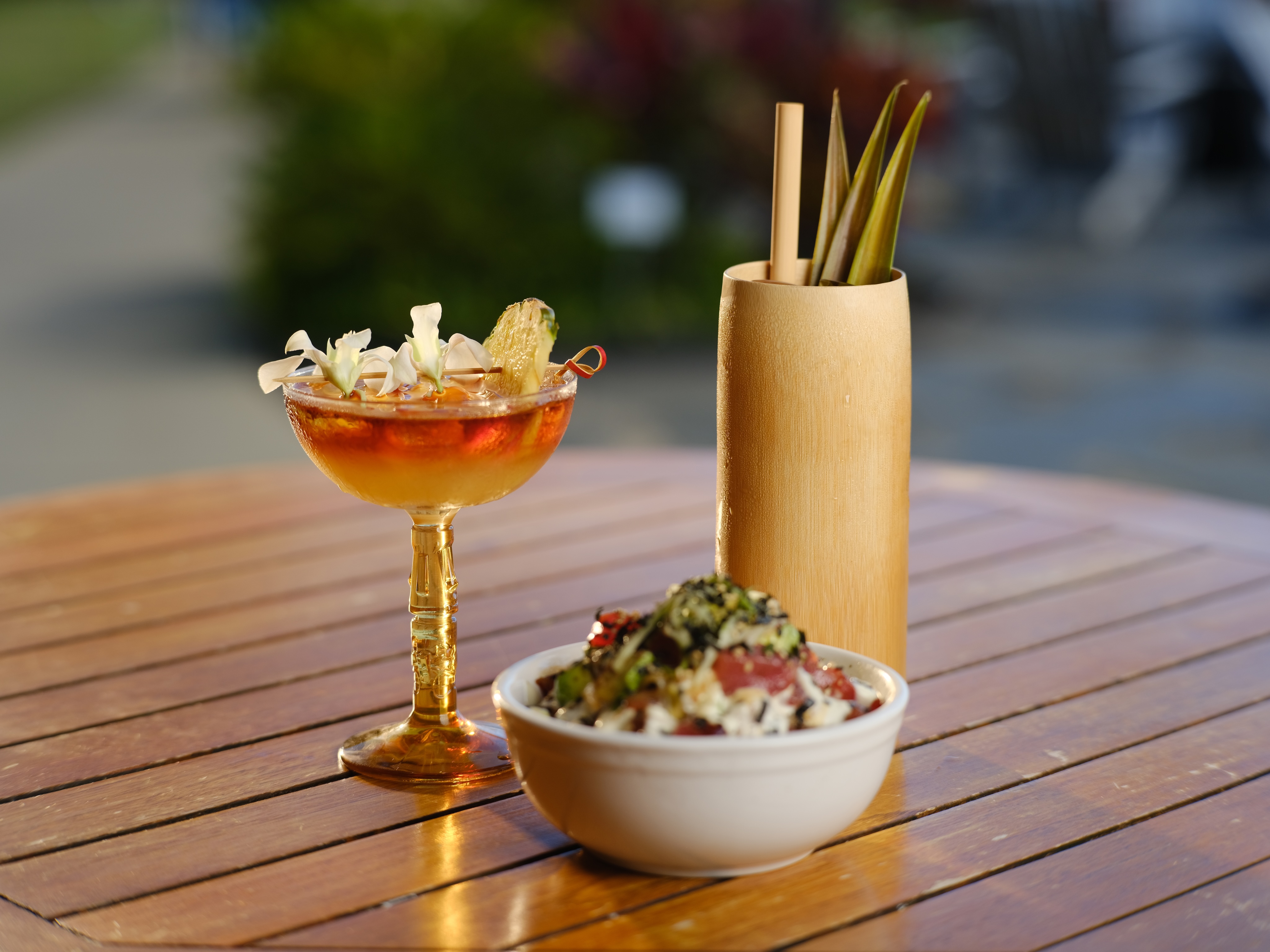 A cocktail with garnishes, a bowl of food with seaweed toppings, and a bamboo container holding sticks are on a wooden table outdoors, setting the scene like an art gallery exhibition in nature.