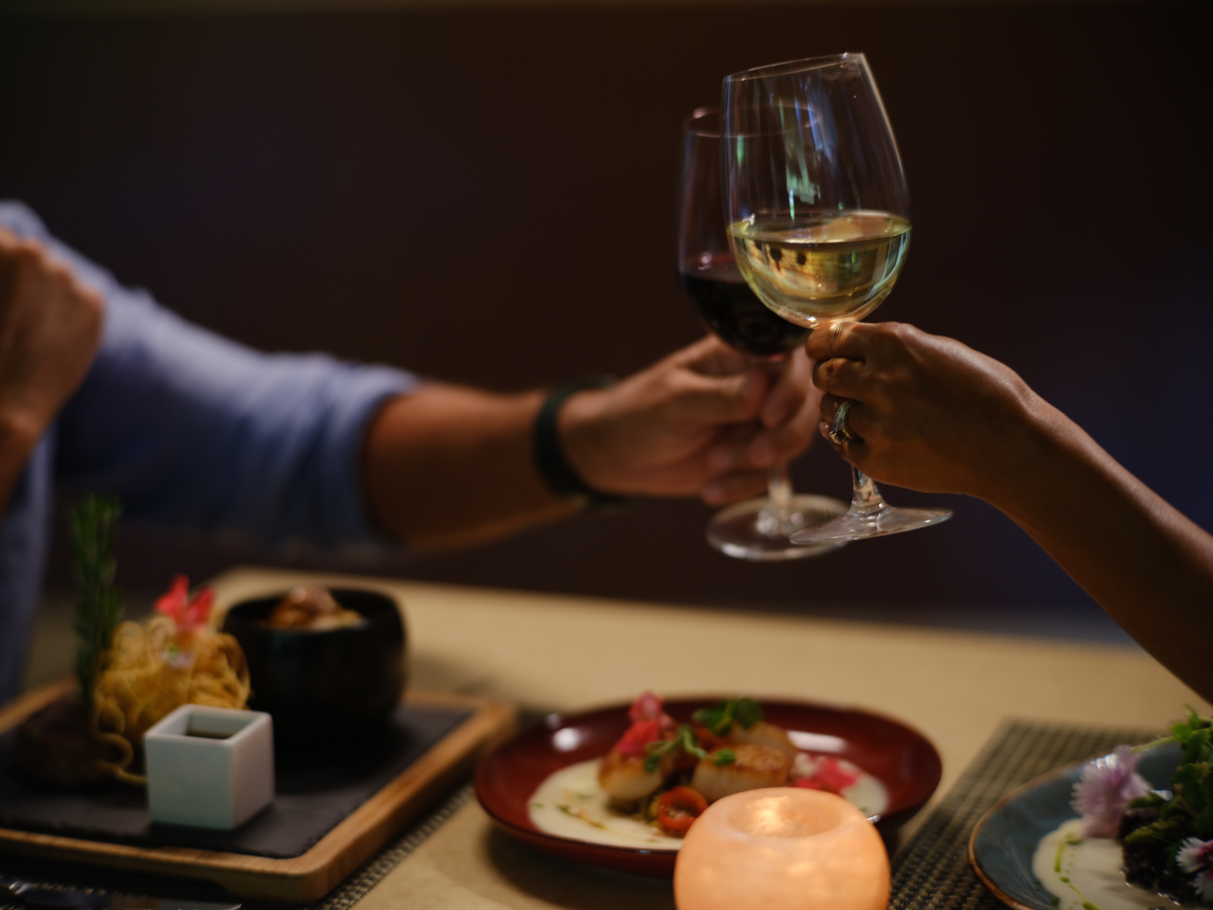 Two people clink red and white wine glasses over a table set with plated meals and a small lit candle in the foreground, creating the perfect setting for intimate private events.