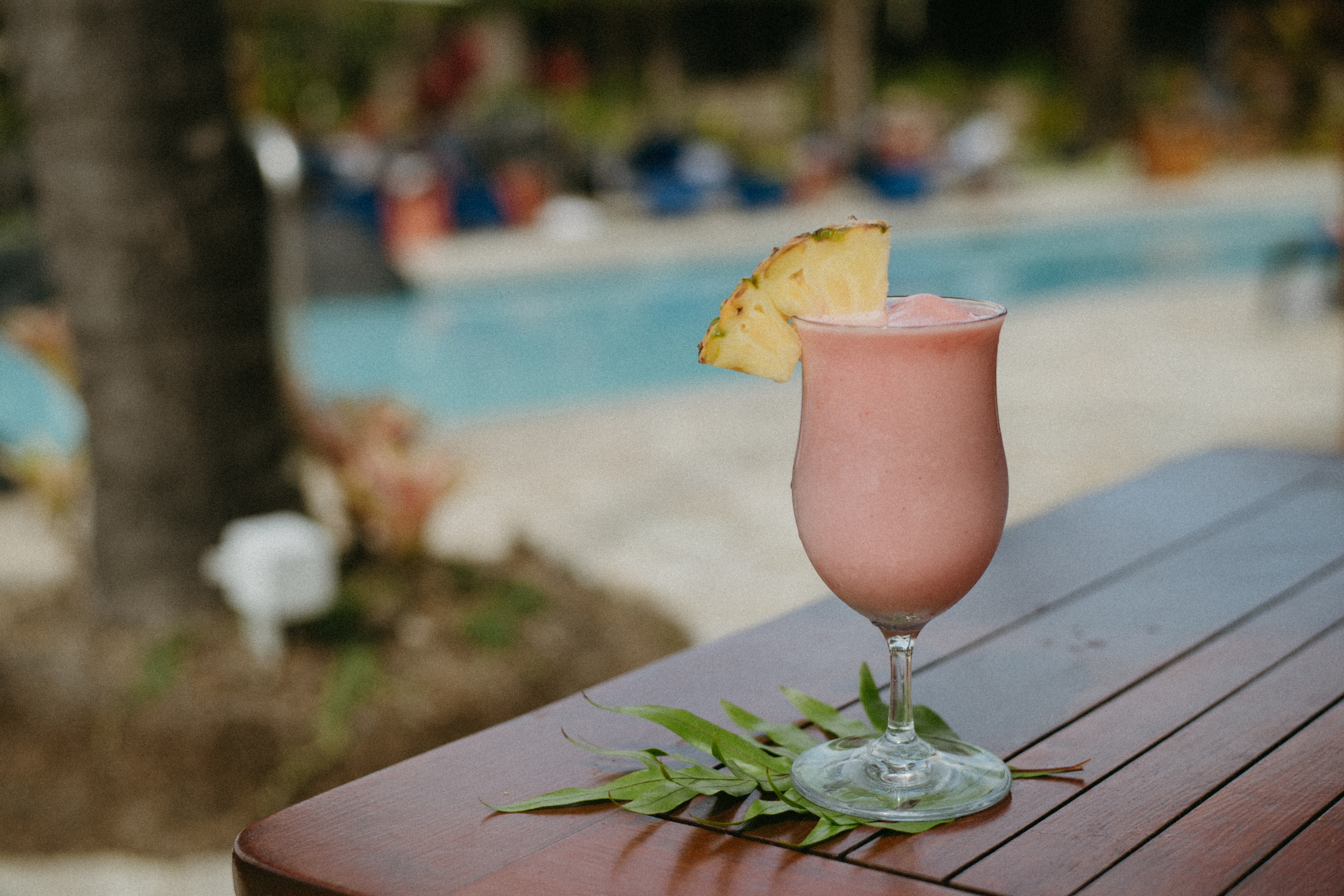 A tropical frozen drink with a pineapple slice garnish sits on a wooden table, reminiscent of an artwork beneath the sun, with a pool and blurry background creating the perfect gallery for summer relaxation.