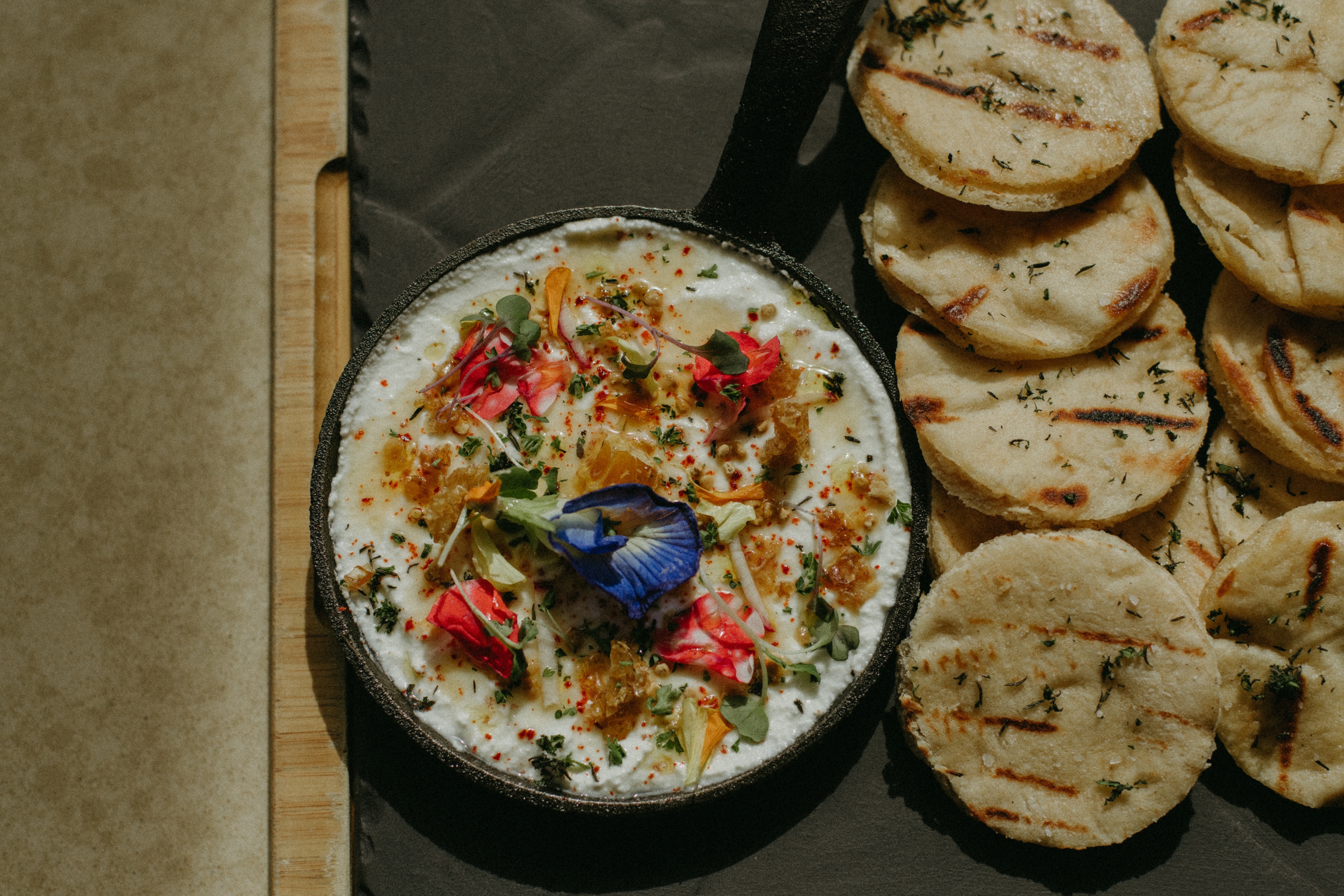 A skillet filled with creamy dip, garnished with edible flowers and herbs, sits like a gallery centerpiece next to a tray of grilled round flatbreads.