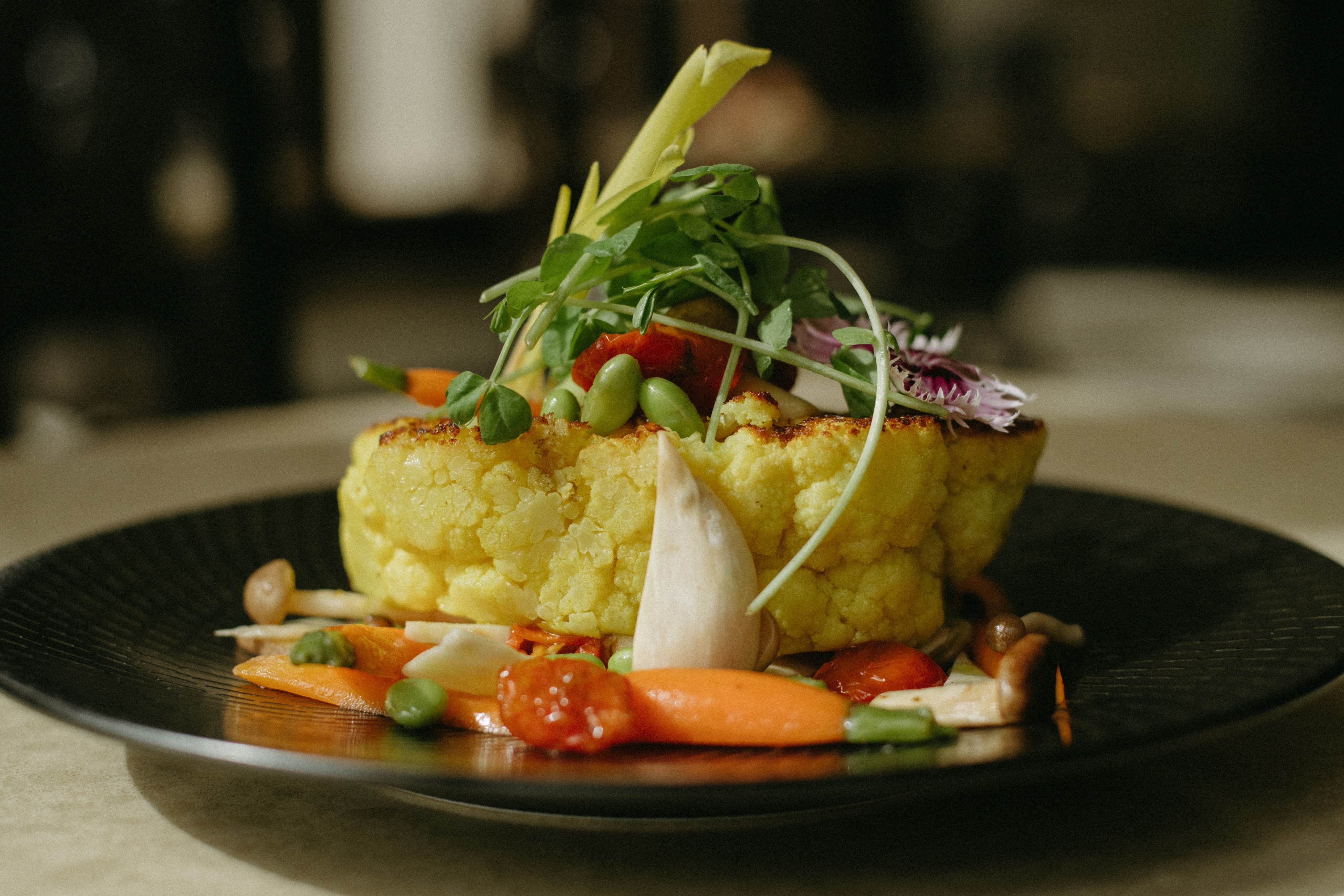 A black plate holds a cauliflower steak garnished with microgreens, served alongside a medley of vegetables—including carrots, edamame, tomatoes, and mushrooms—like an art exhibition on display.