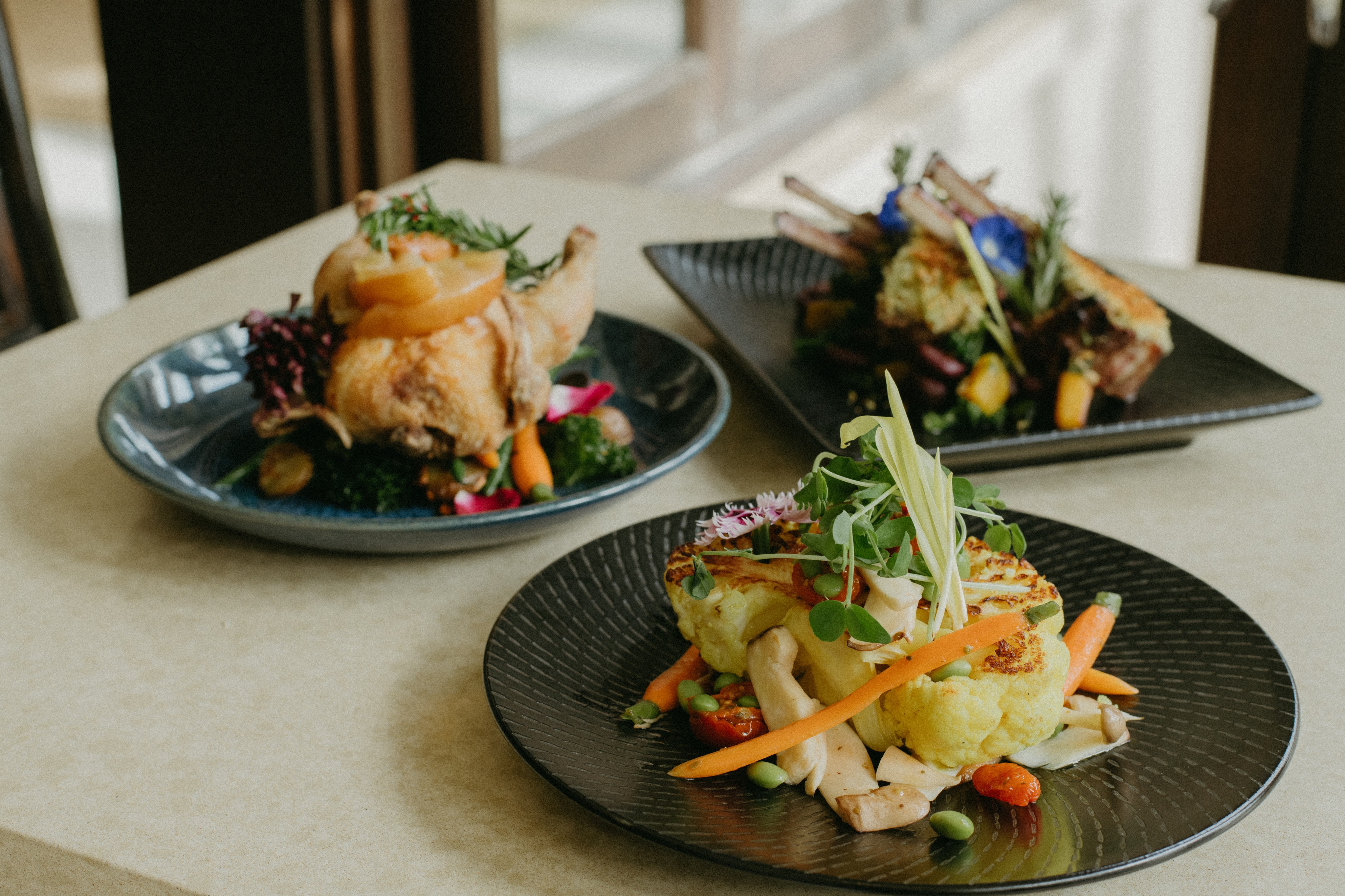 Overview: Three elegantly plated dishes on a table: roasted chicken with vegetables, rack of lamb with garnish, and a vegetarian patty with vegetables and microgreens.