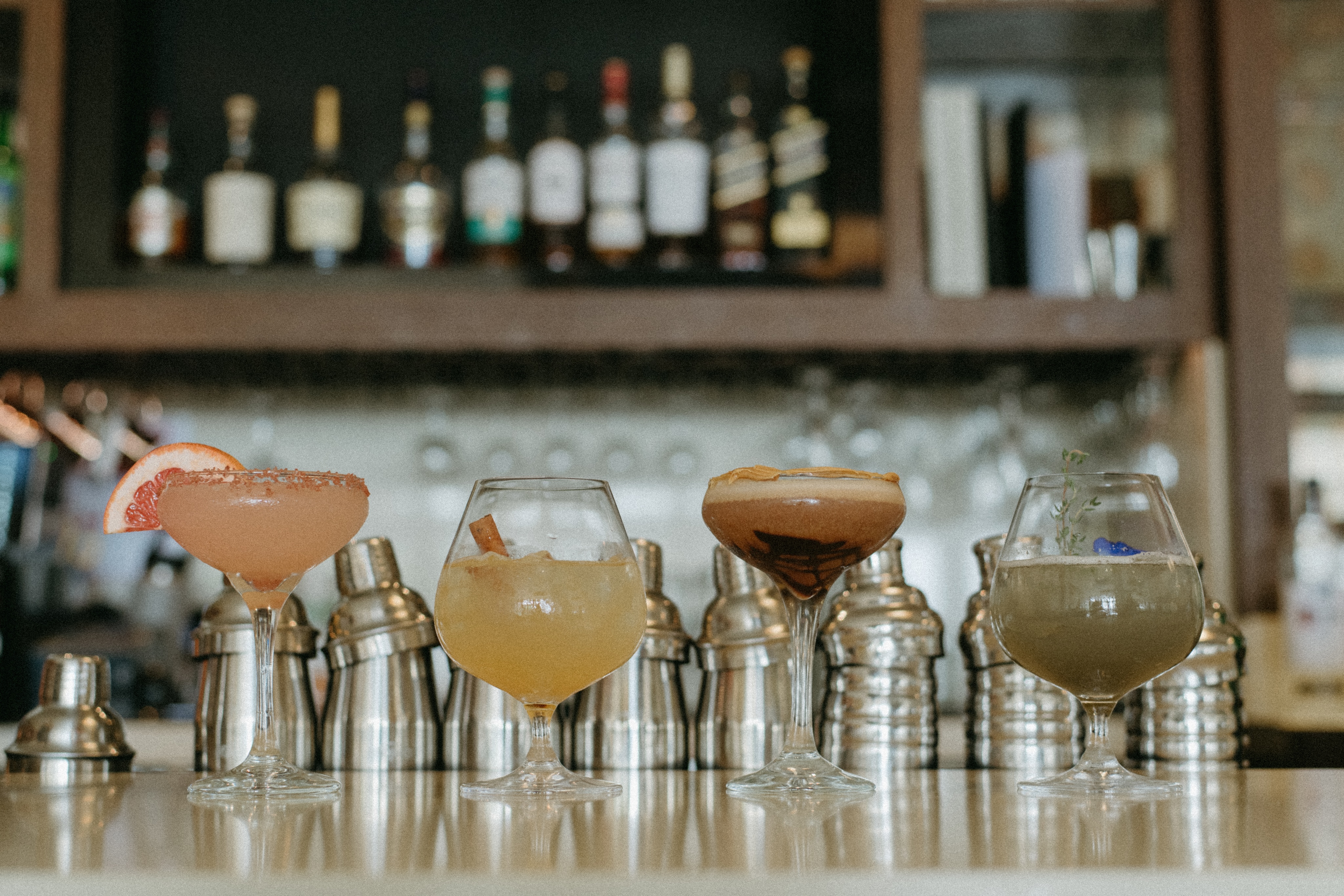 Four cocktails, each in a distinct glass, are lined up on a bar counter resembling an art gallery exhibition, with cocktail shakers and a shelf of bottles completing the captivating display.