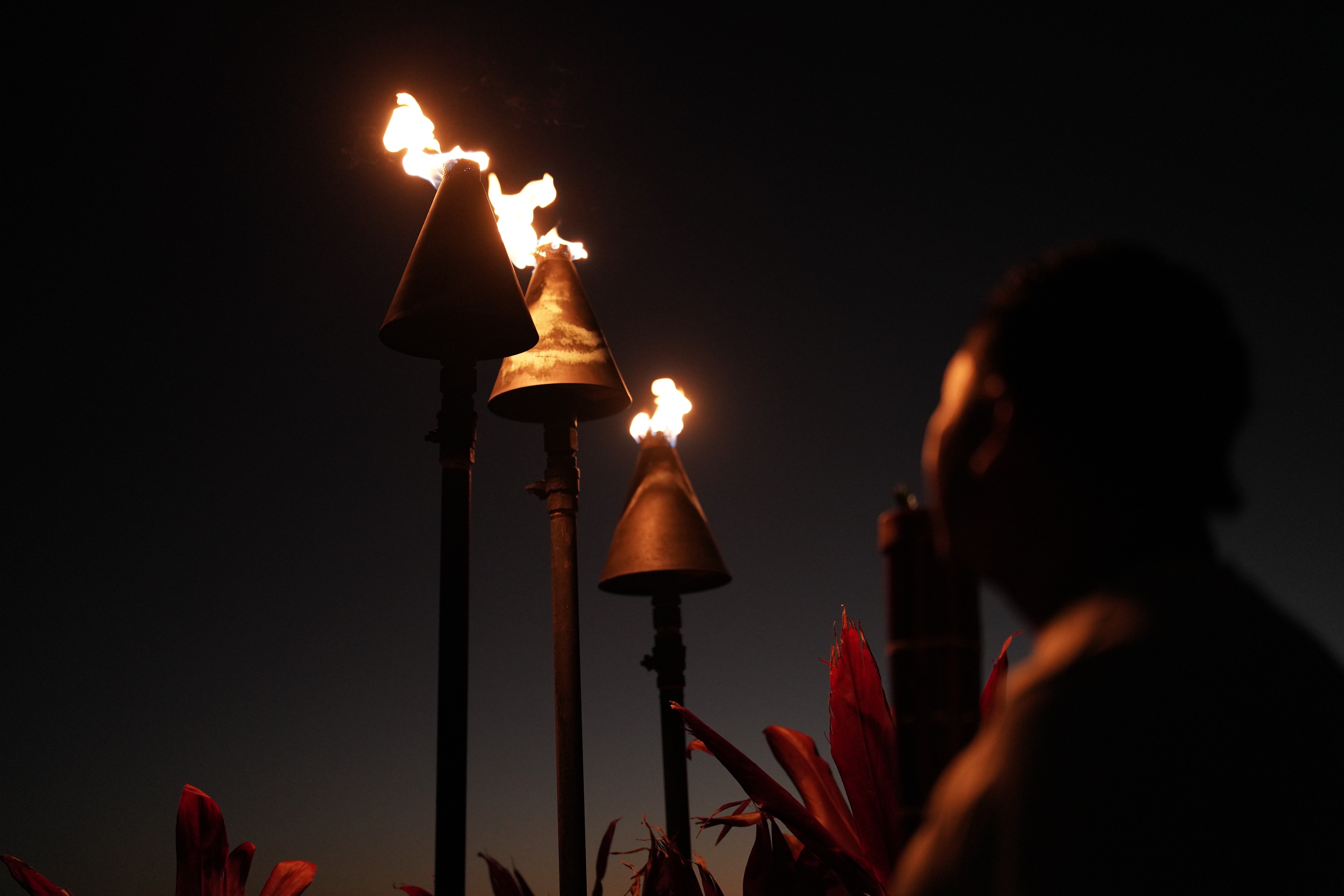 Silhouette of a person looking at three lit tiki torches against a dark sky, surrounded by red foliage, creating a captivating image perfect for the homepage.