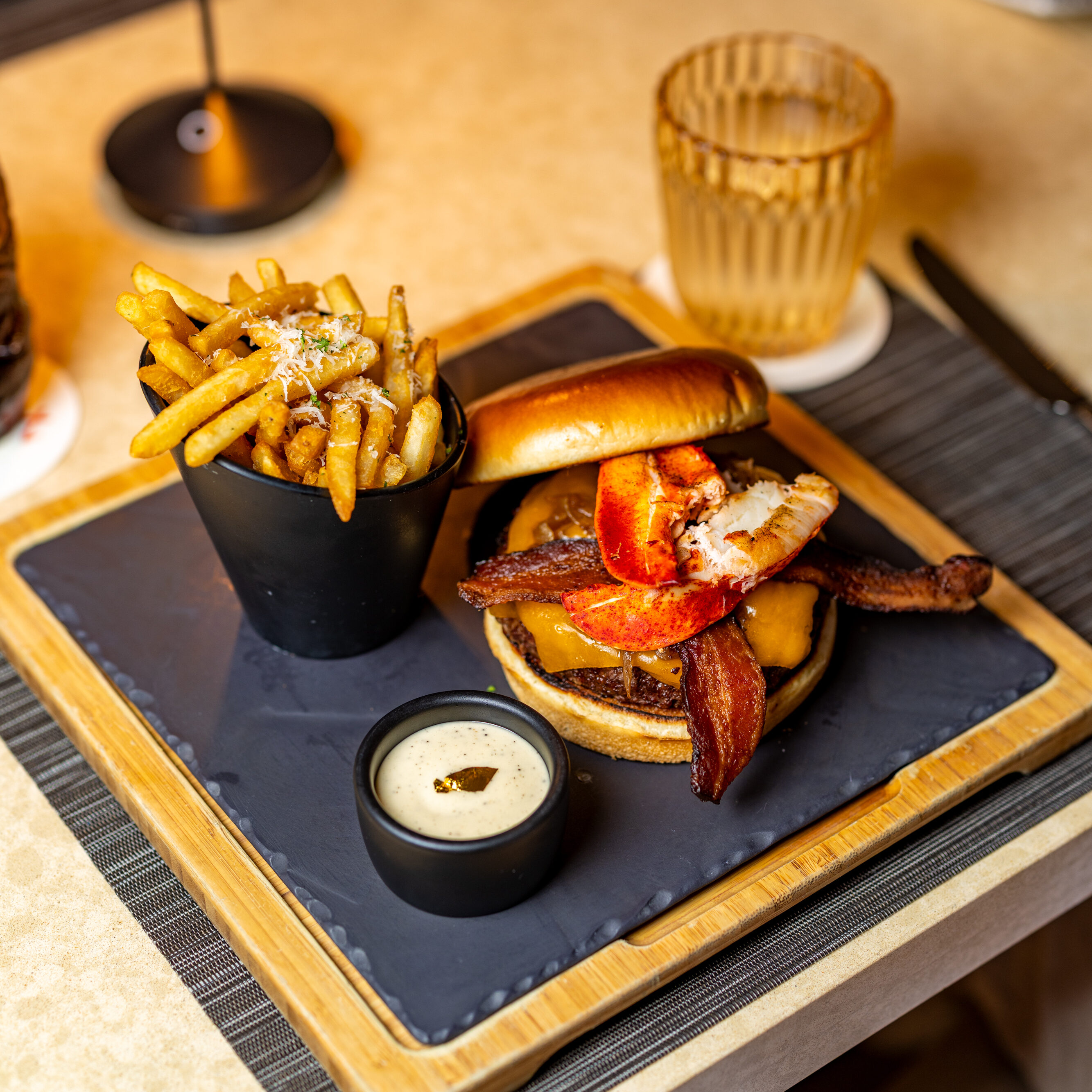 A burger topped with bacon and lobster is served with a side of fries and dipping sauce on a square plate, next to a glass of iced drink on a beige table.