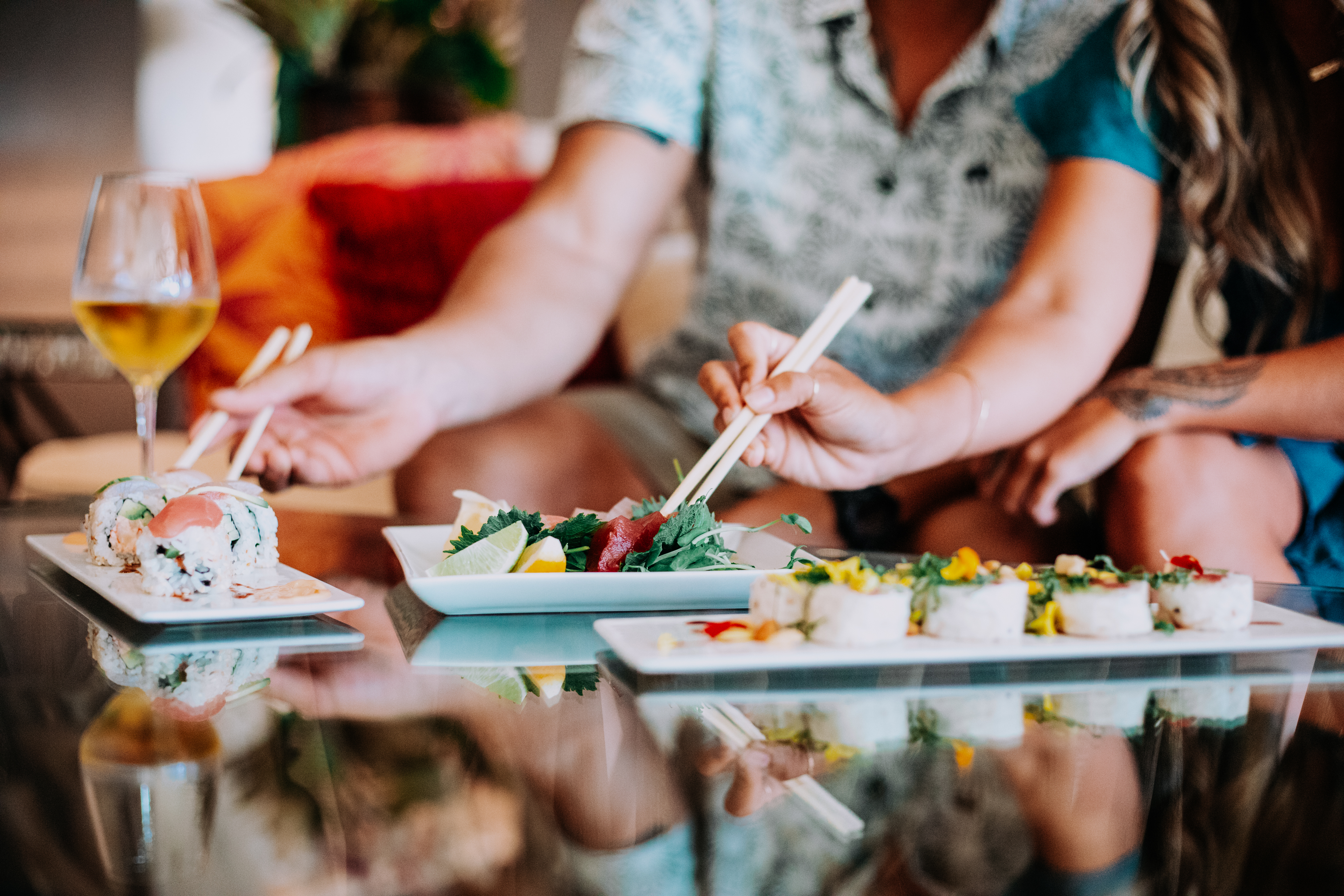 Two people are dining at home, using chopsticks to eat sushi and a green salad from white plates, with a glass of white wine visible on the table.