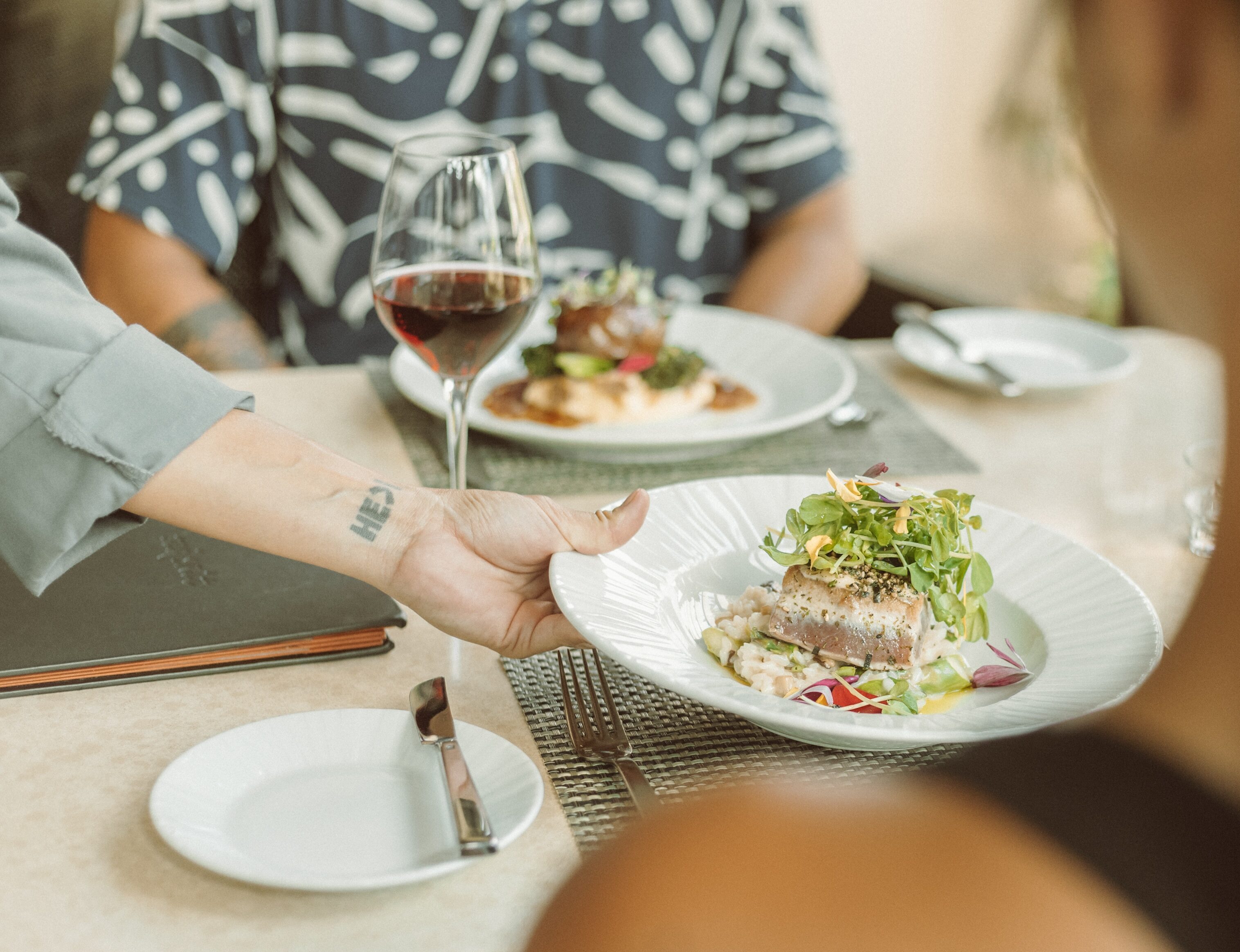 A server hands a dish of cooked fish topped with greens to a seated person at a restaurant table set with wine, another plate of food, and dining utensils. This vivid description captures the inviting ambiance of fine dining.