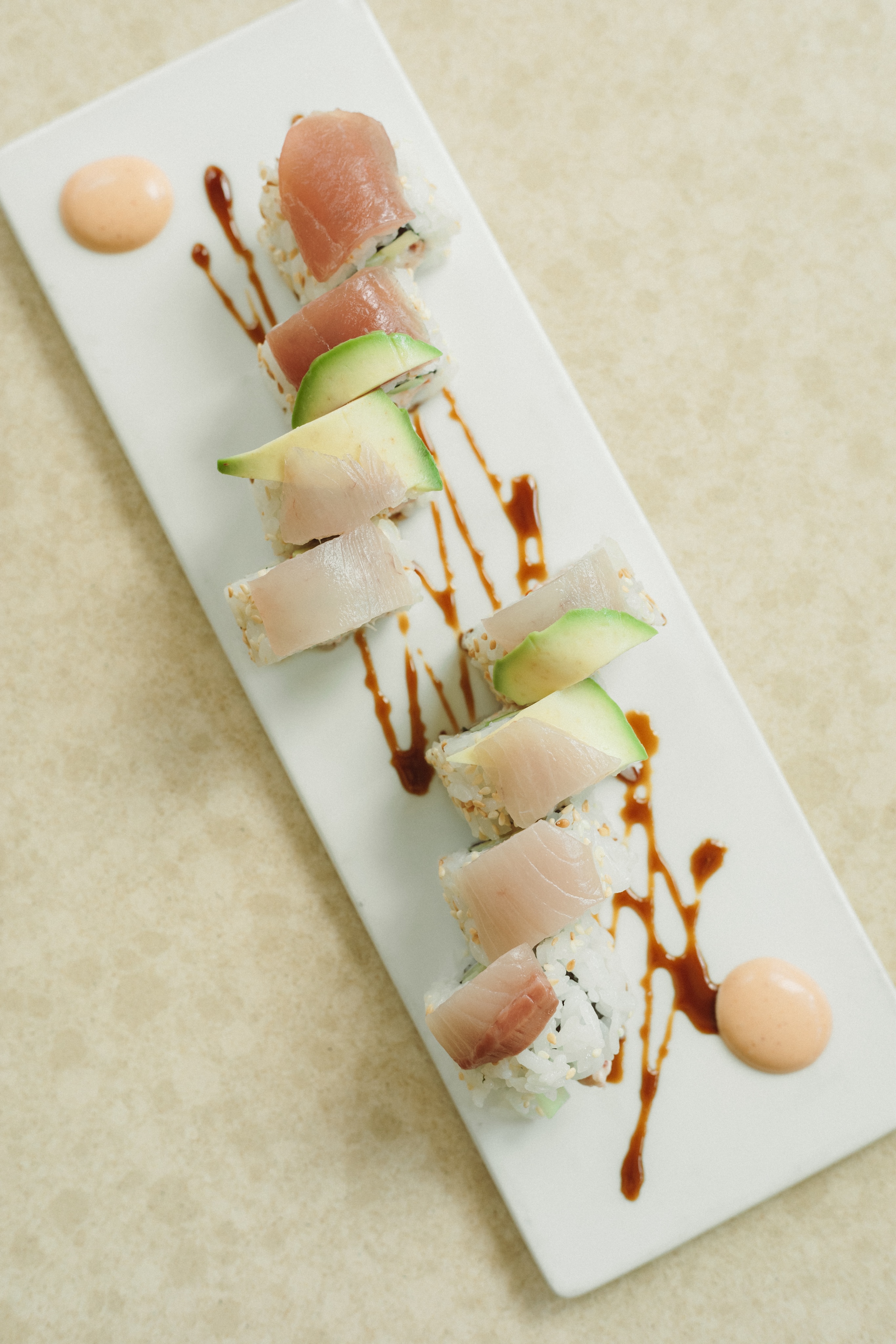 A rectangular white plate with sushi rolls topped with fish and avocado, drizzled with sauce, and accompanied by two dollops of orange-colored sauce on a light surface, resembling an art piece in a gallery.