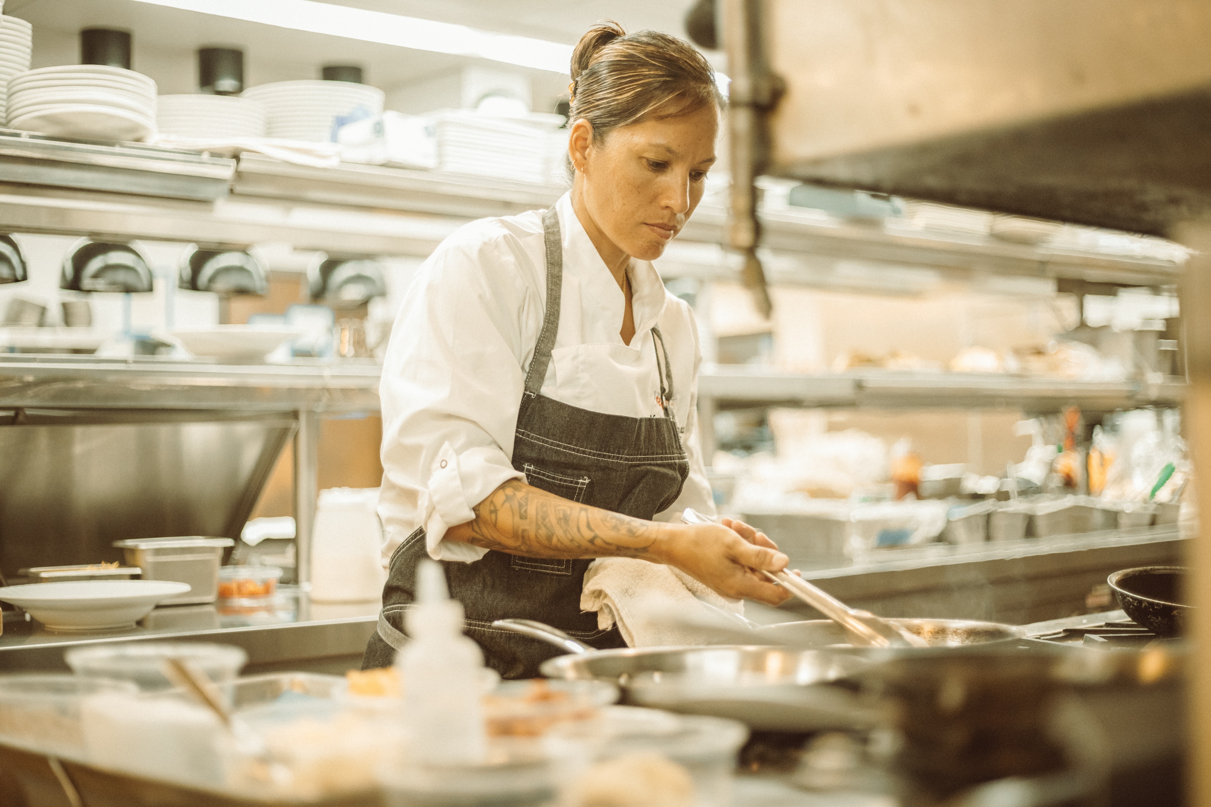 A chef in a white shirt and gray apron is focused on cooking in a commercial kitchen, surrounded by cooking utensils and ingredients about the space.