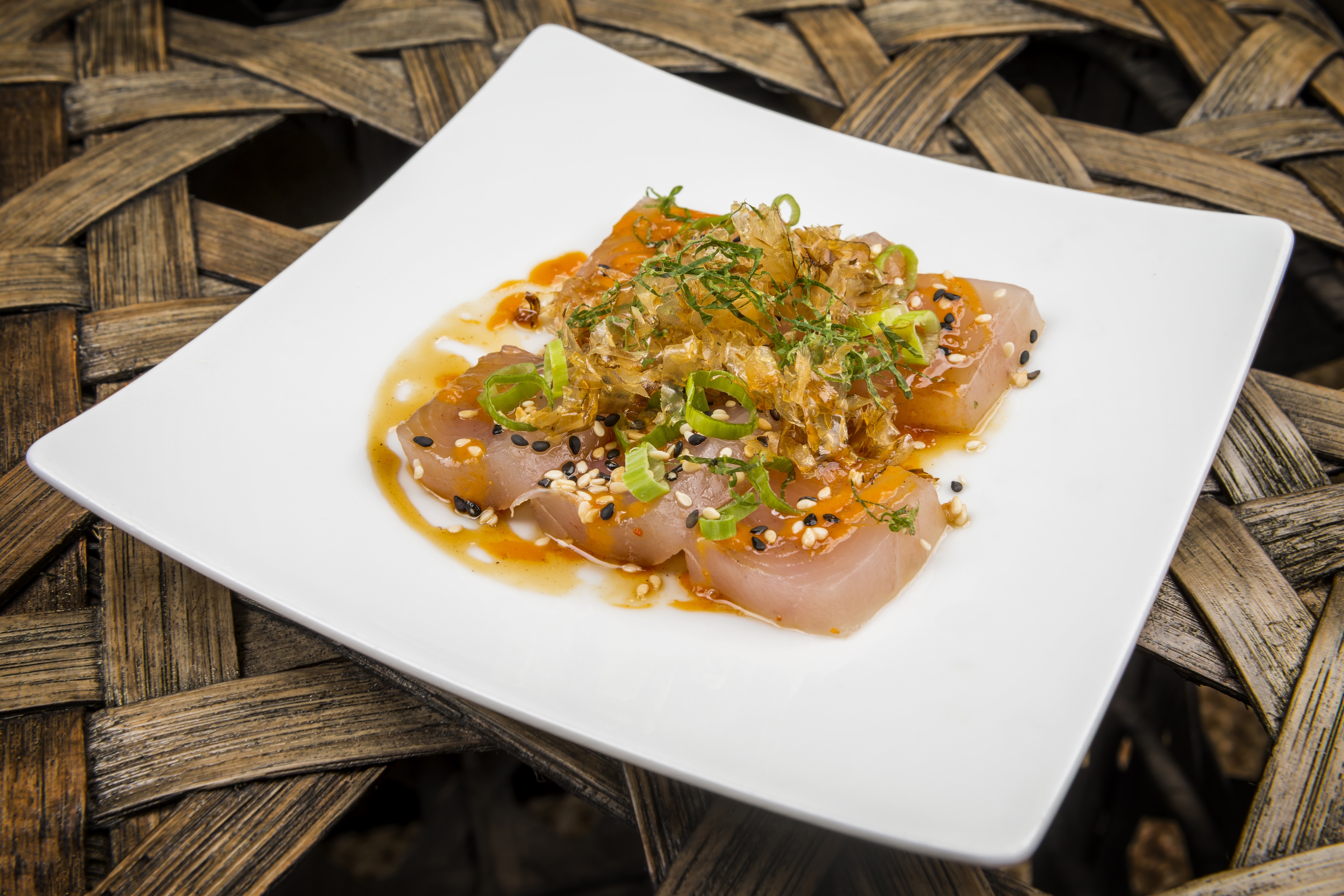 A white square plate with thinly sliced raw fish topped with green onions, sesame seeds, and a light sauce, elegantly displayed like a culinary gallery piece on a wicker table.