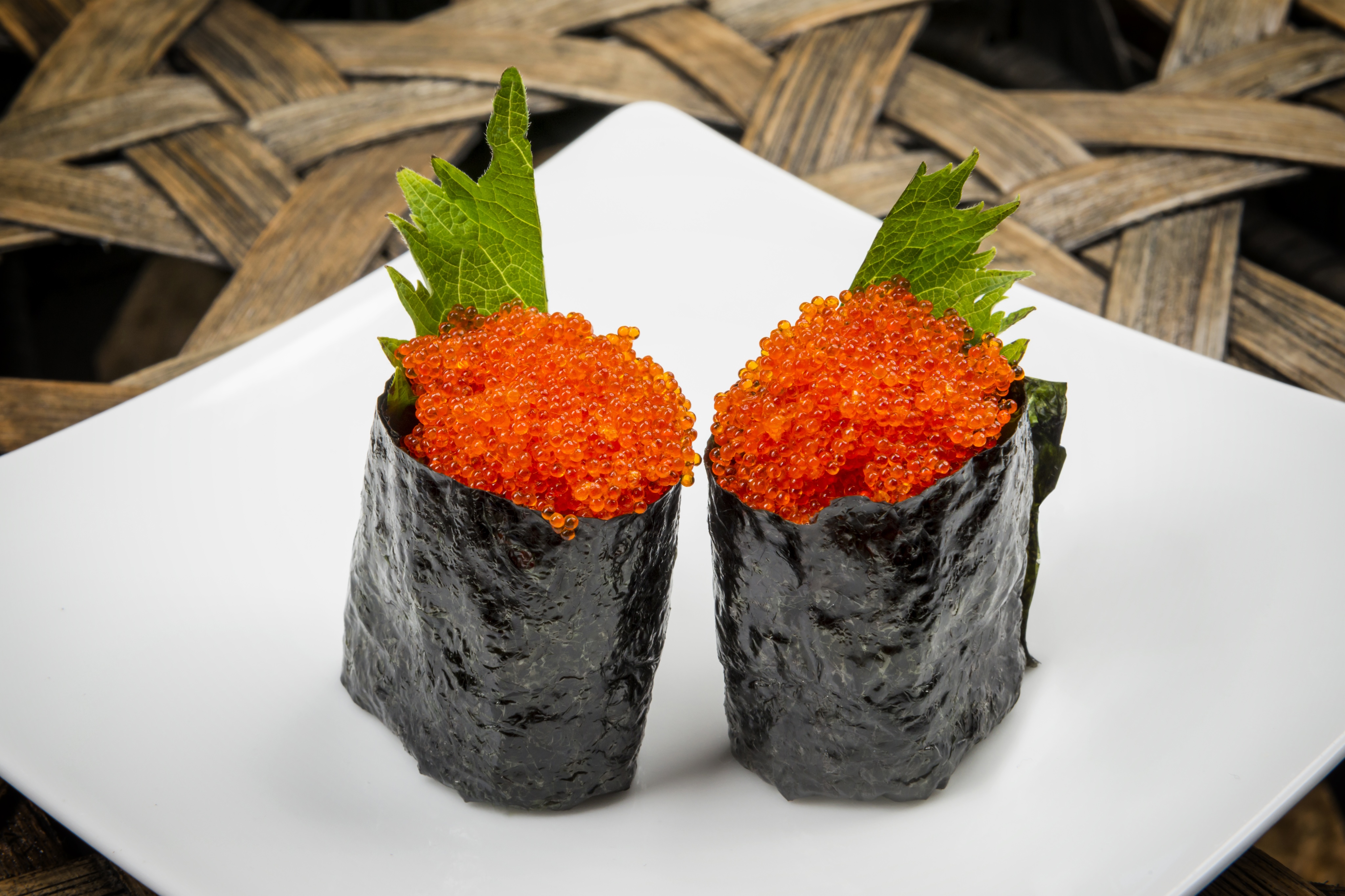 Two pieces of sushi, garnished with orange fish roe and green leaves, wrapped in seaweed, are artfully arranged on a white square plate. The presentation resembles a culinary gallery masterpiece.