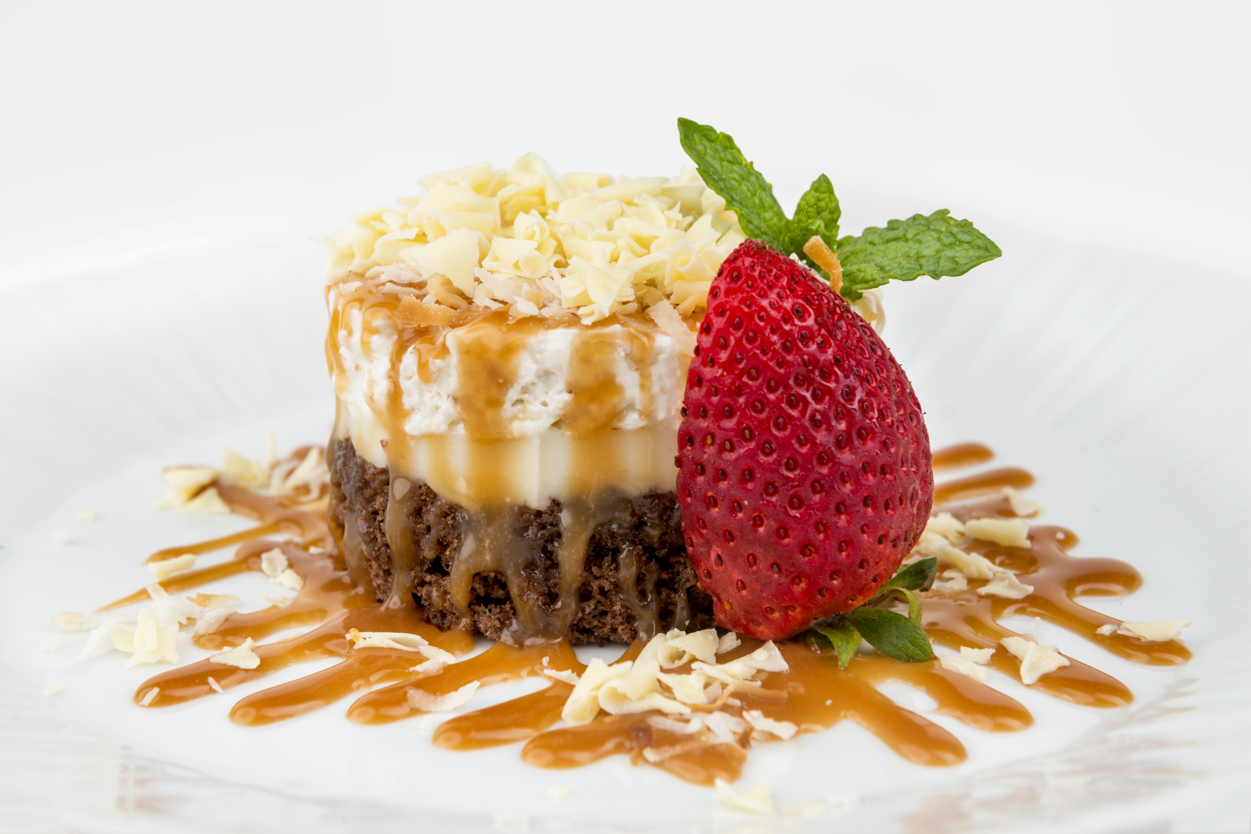 A small layered dessert with chocolate and caramel sauce, topped with white chocolate shavings, a strawberry, and mint leaves on a white plate, perfect for an art gallery exhibition.