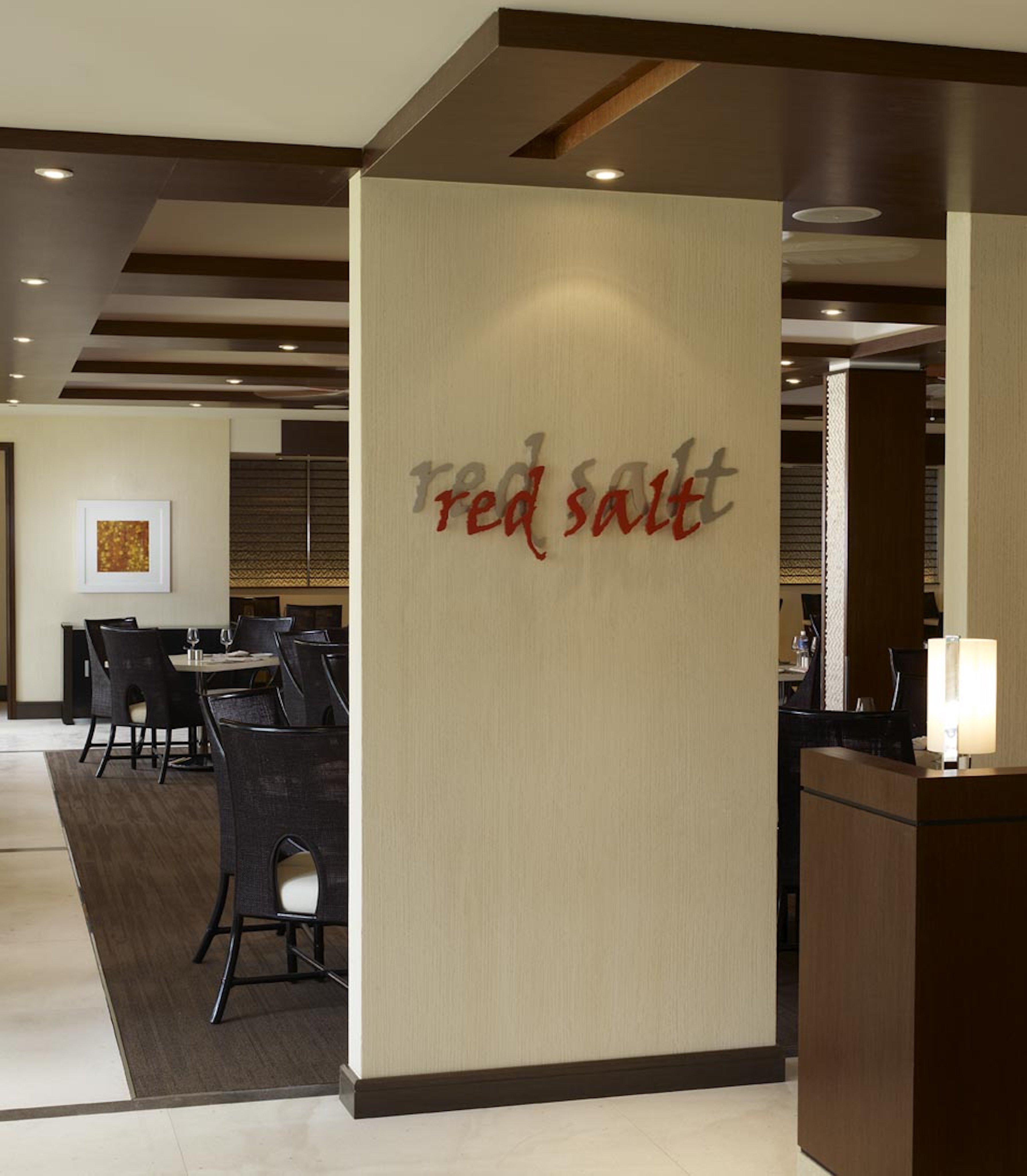 Interior view of a restaurant with a sign reading "red salt" on a wall. The space is furnished with tables and chairs under a ceiling with recessed lighting, resembling an elegant gallery. Artwork is visible on the far wall.