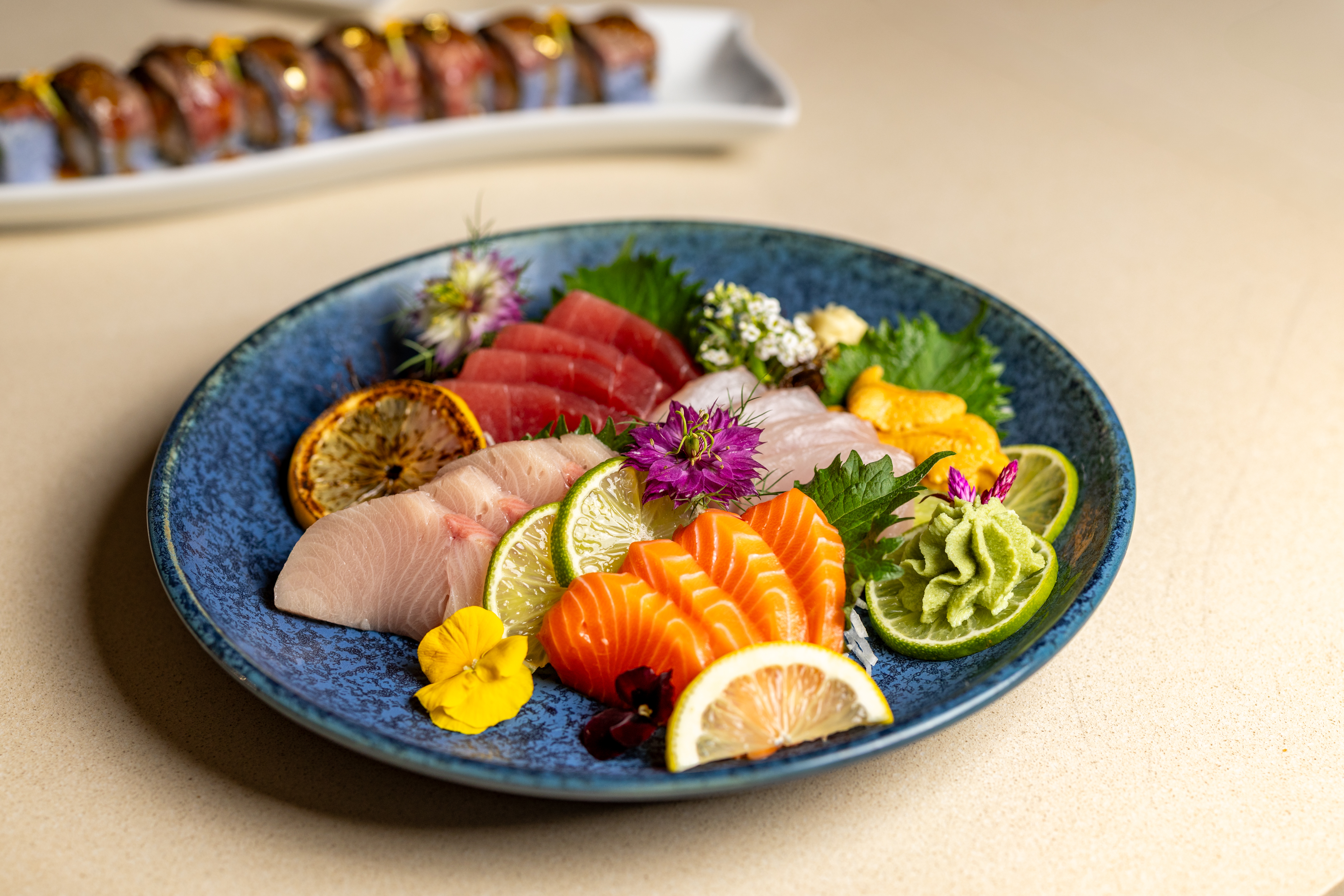 A blue plate with assorted sashimi, garnished with lime, lemon, edible flowers, and wasabi; sushi rolls are blurred in the background.