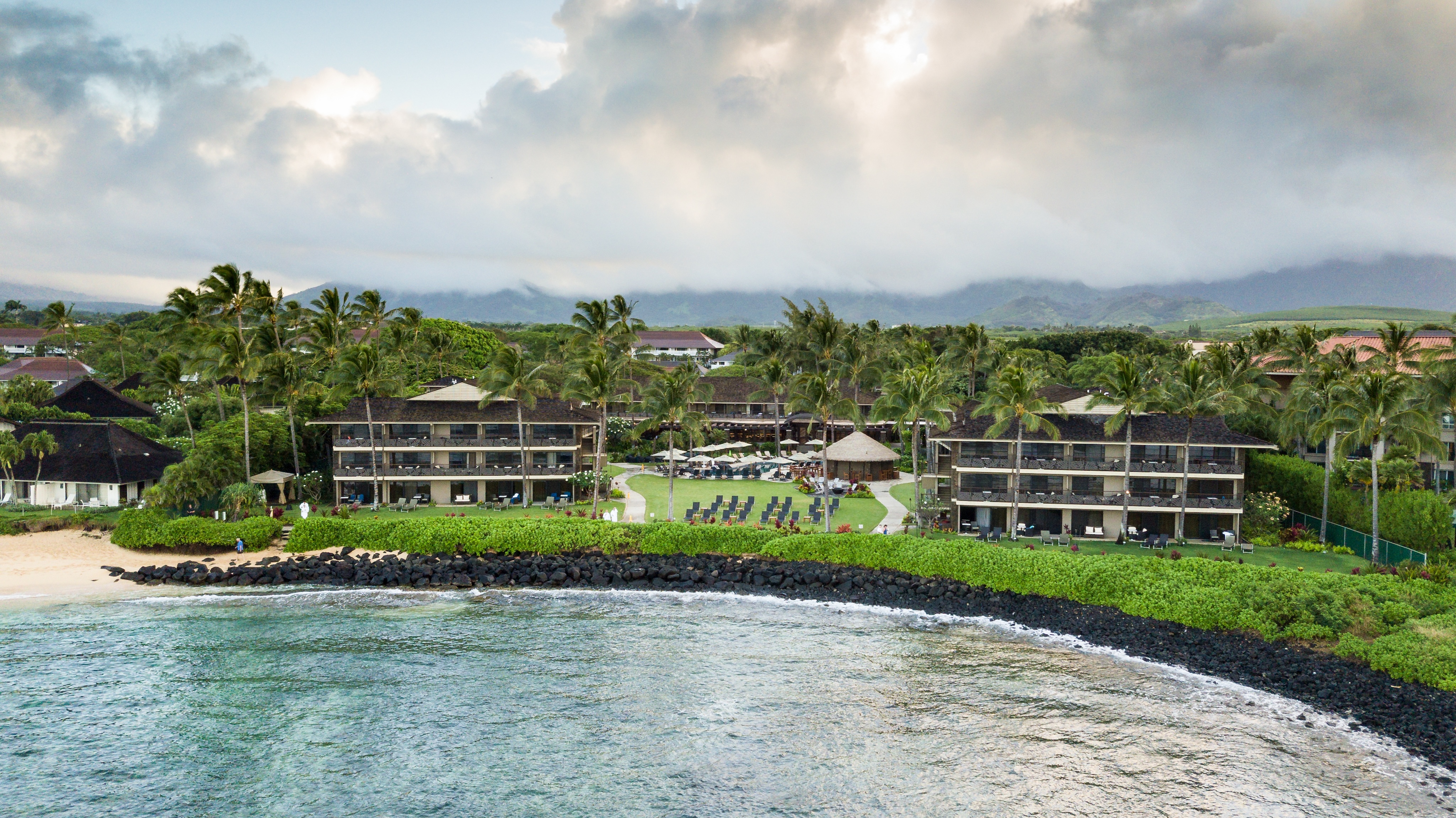 Aerial view of a beachfront resort with several buildings, palm trees, lounge chairs on a lawn, and rocky shoreline under a cloudy sky, reminiscent of an art gallery showcasing nature's masterpieces.