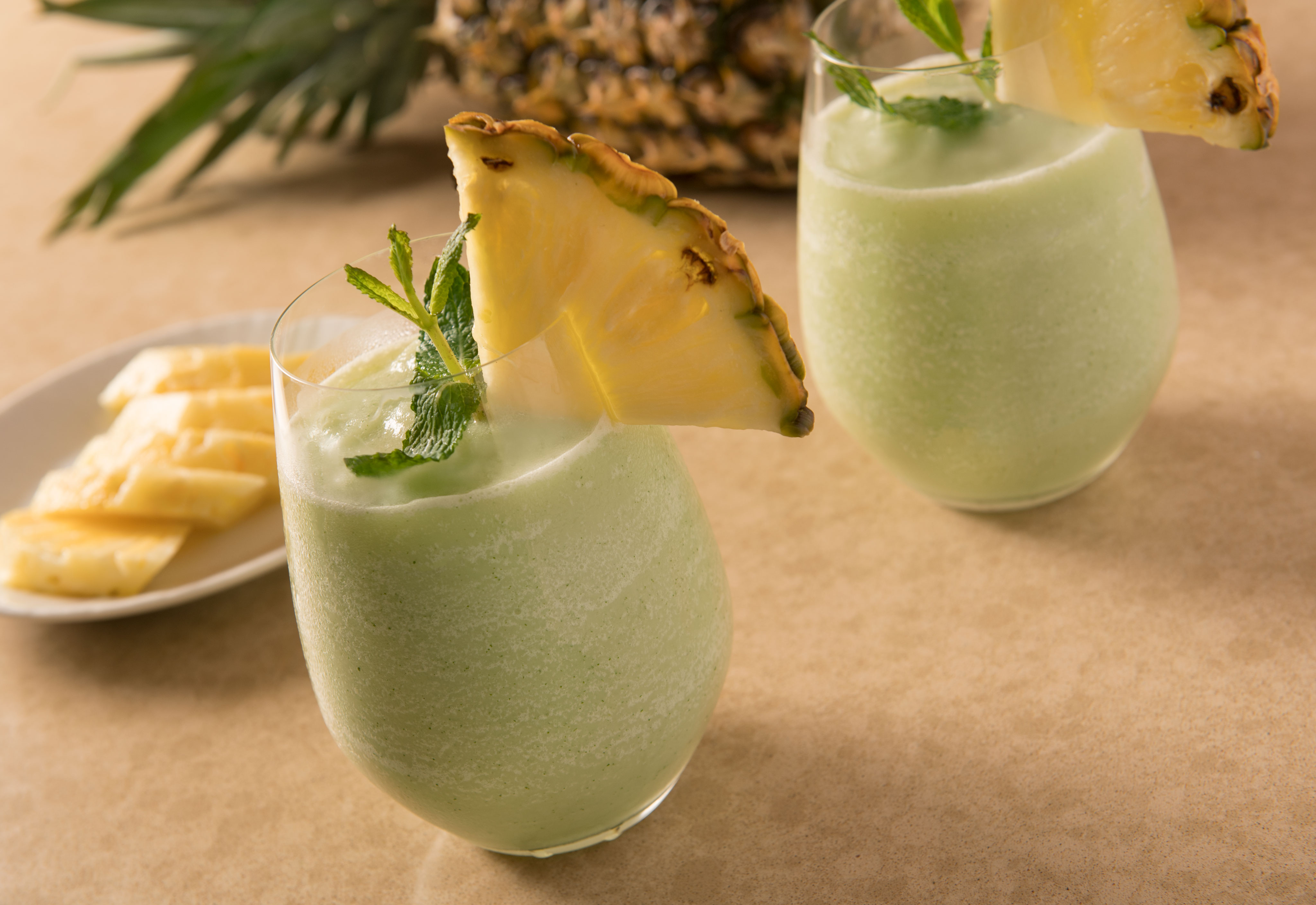 Two glasses of green smoothies garnished with pineapple slices and mint leaves, set on a light-colored surface like a curated artwork in an exhibition, with a plate of pineapple chunks and a whole pineapple in the background.