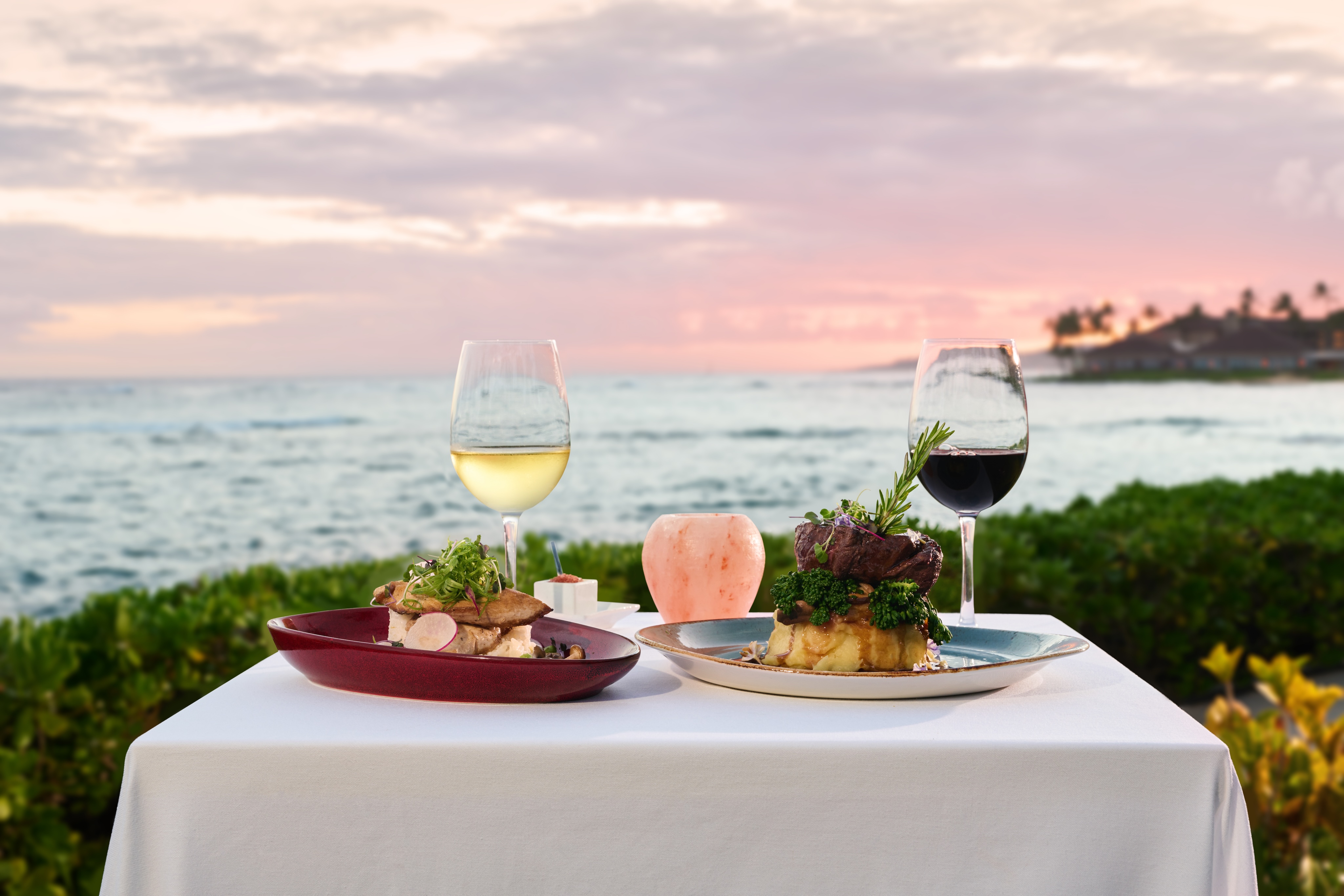 A private cabana dinner for two overlooking the ocean at sunset, featuring two glasses of wine and two plates of gourmet food on a white tablecloth.