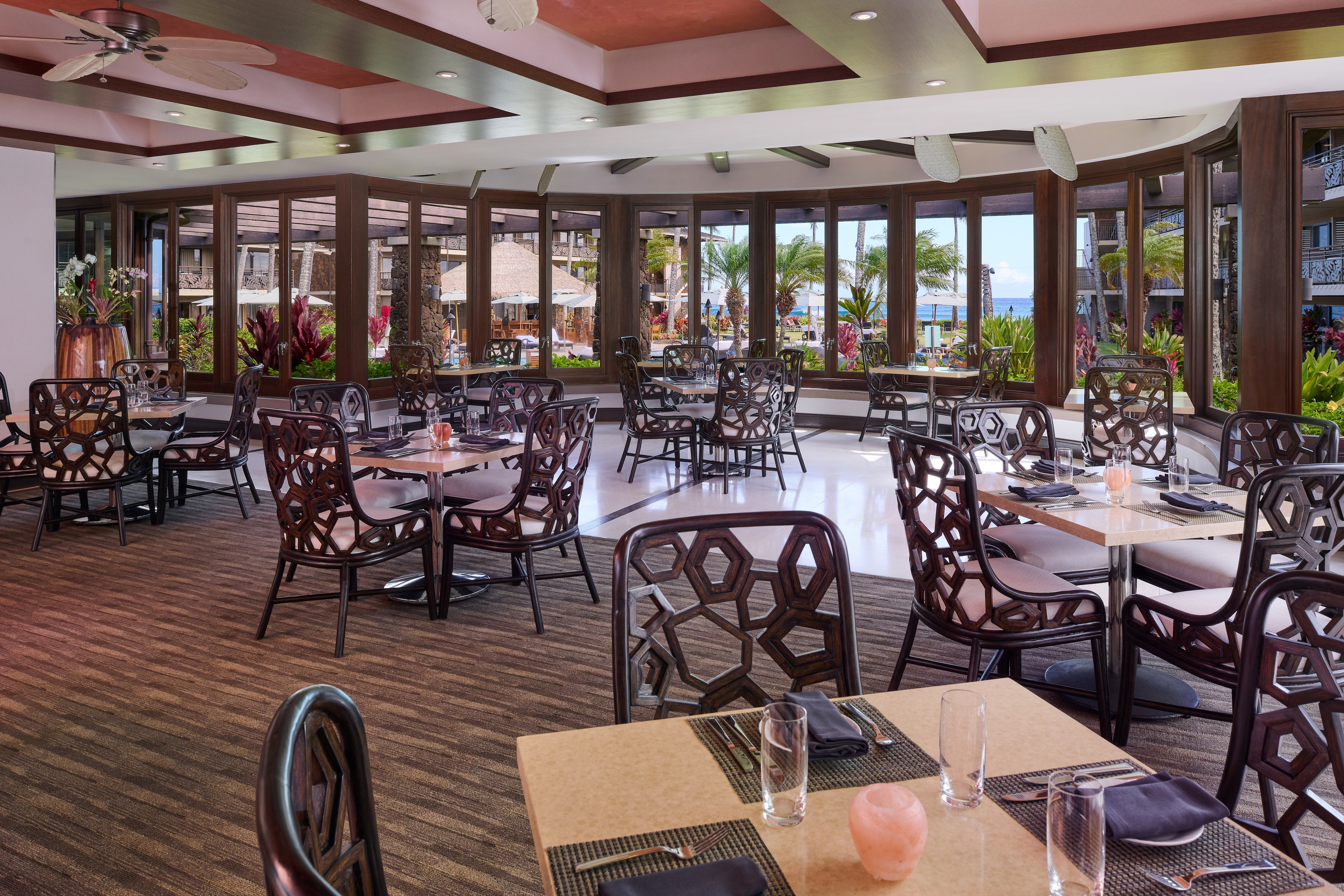 Restaurant dining area with modern decor, featuring patterned chairs, tables set with menus and glassware, large windows providing natural light and views of outdoor greenery and a pool area.