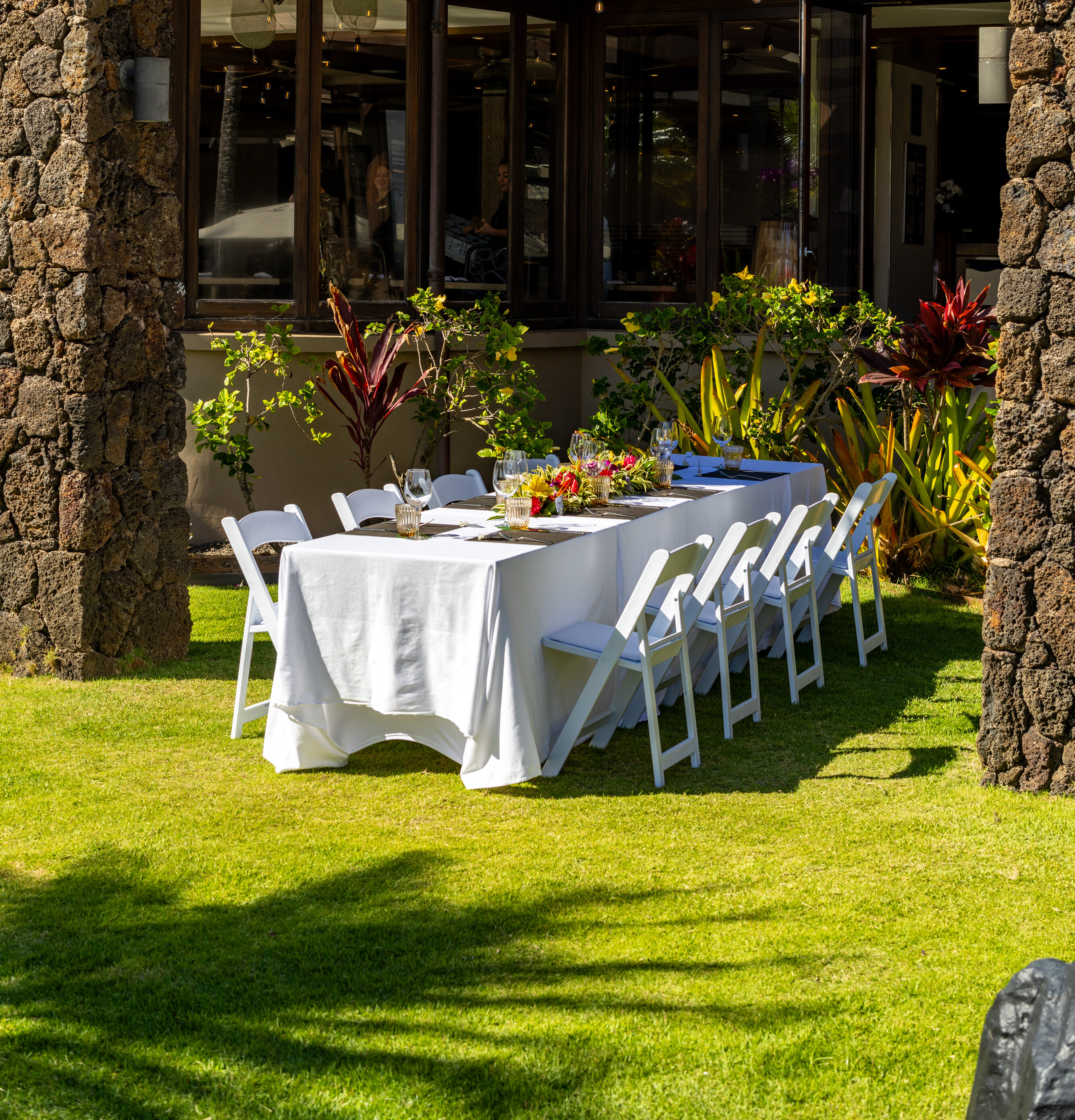 A long outdoor dining table with a white tablecloth and white chairs is set for a meal on a lawn under a pergola, creating a lanai-inspired setting next to a stone building, perfect for savoring dishes seasoned with Red Salt.