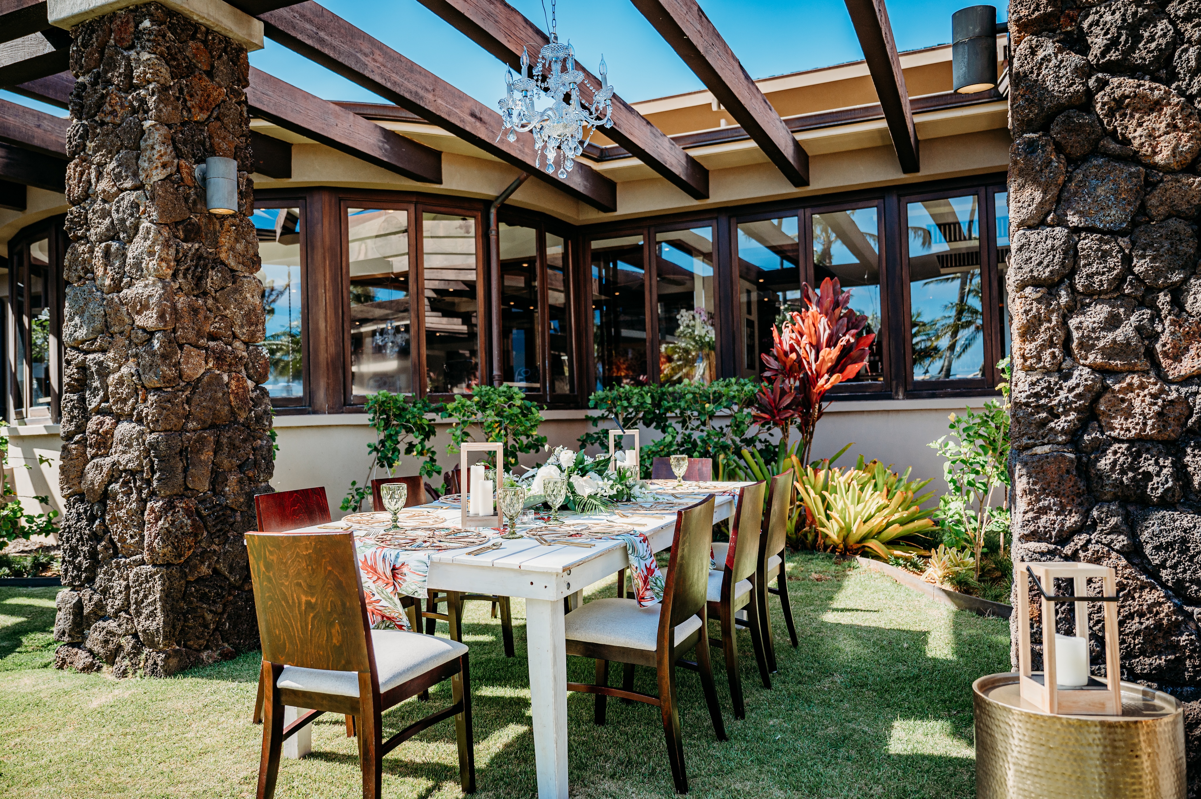 An outdoor dining table with six chairs set up under a wooden pergola with a chandelier, surrounded by stone pillars and tropical plants, perfect for hosting intimate private events.