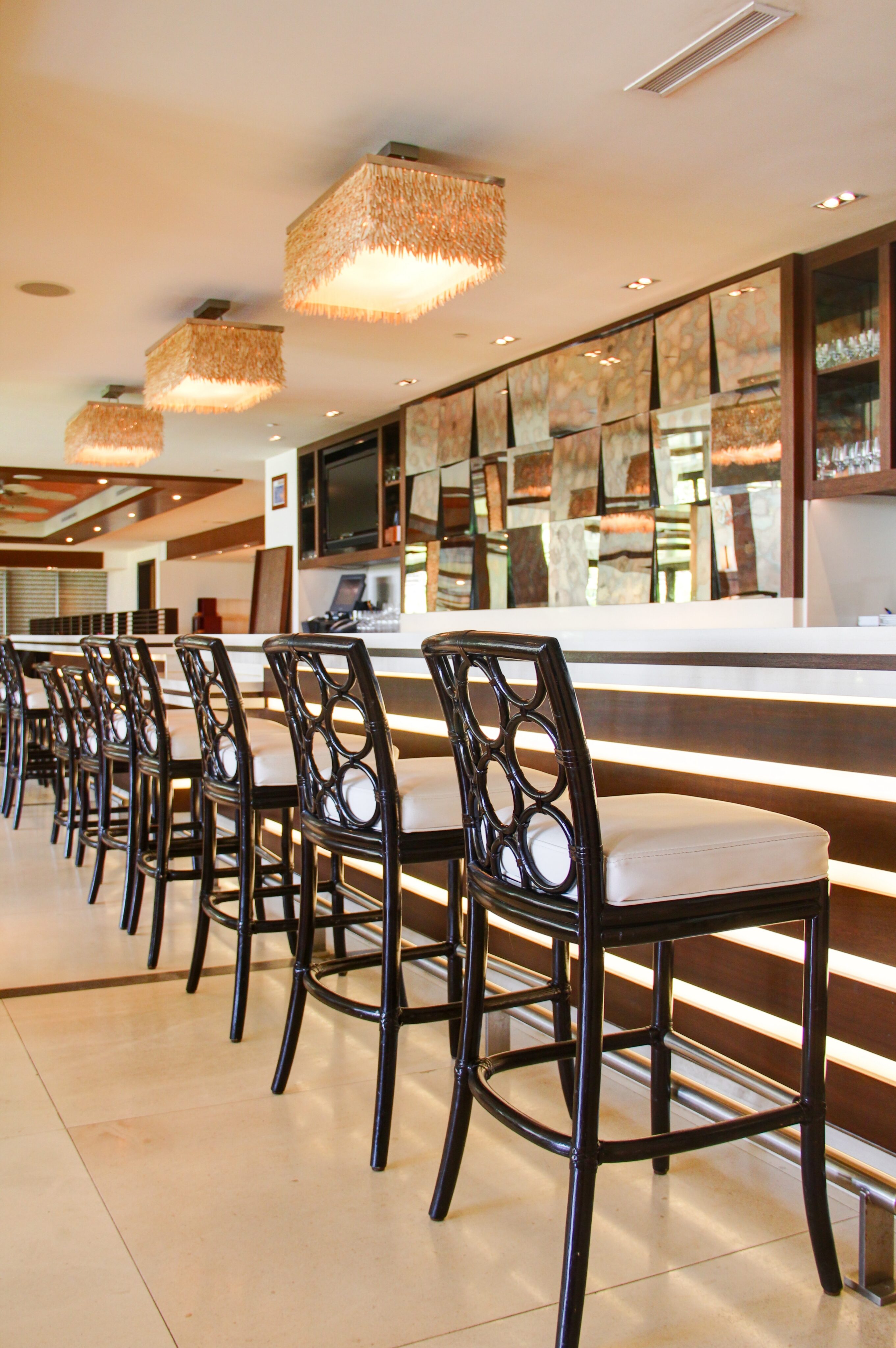 Modern bar interior with high chairs lined up along a counter, illuminated by stylish hanging lights and decorated with mirrored panels on the wall, evoking a chic gallery ambiance.