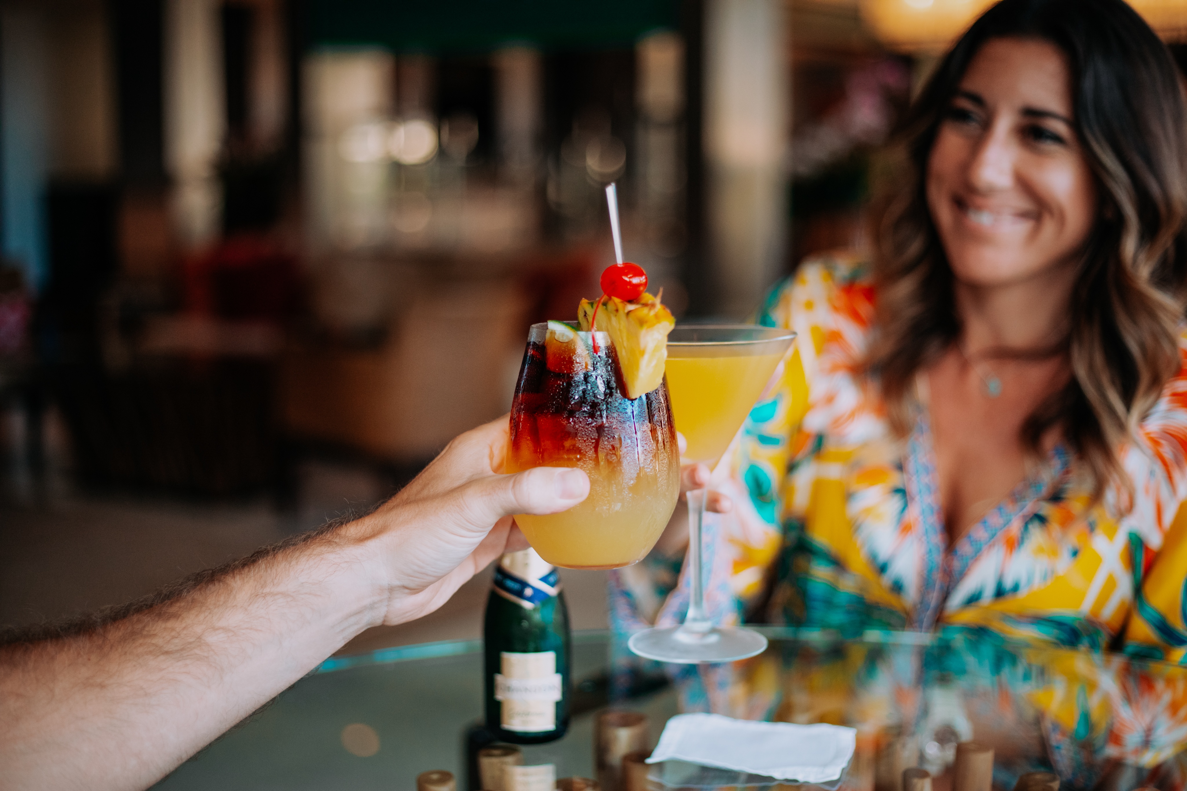 Two people, one shown partly, cheers with colorful cocktails at a glass table. One drink has a cherry and pineapple garnish, and a small bottle is seen on the table. The setting feels like an exclusive art gallery with its elegant yet blurred indoor background.