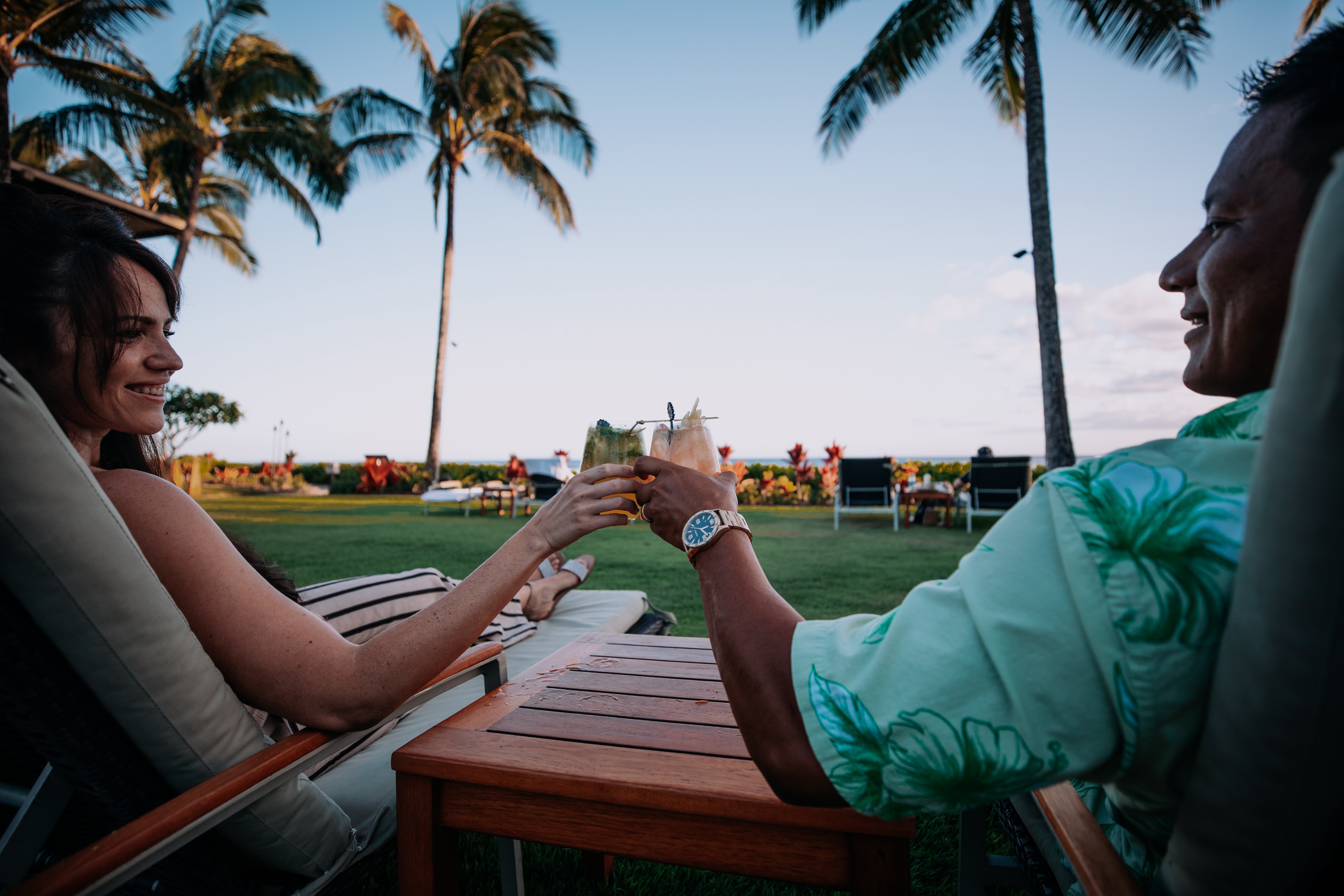 Two people relax on lounge chairs near a lawn and palm trees, clinking glasses of drinks against a backdrop of a clear sky and distant water.