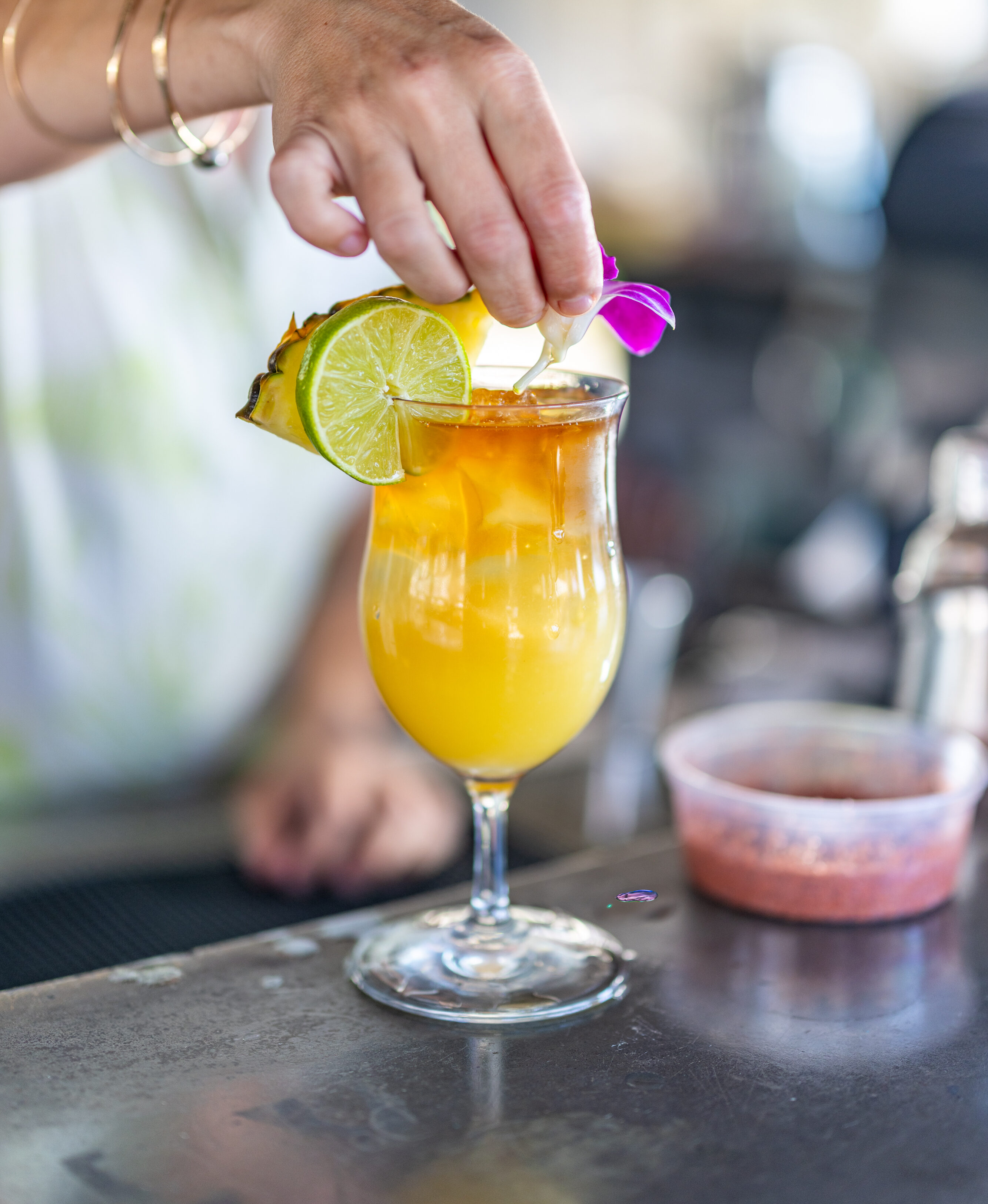 A person garnishes a yellow tropical cocktail in a stemmed glass with a slice of lime, pineapple, and a purple flower, perfect for serving at a Manager's Meet or elegant Meet and Greet event.