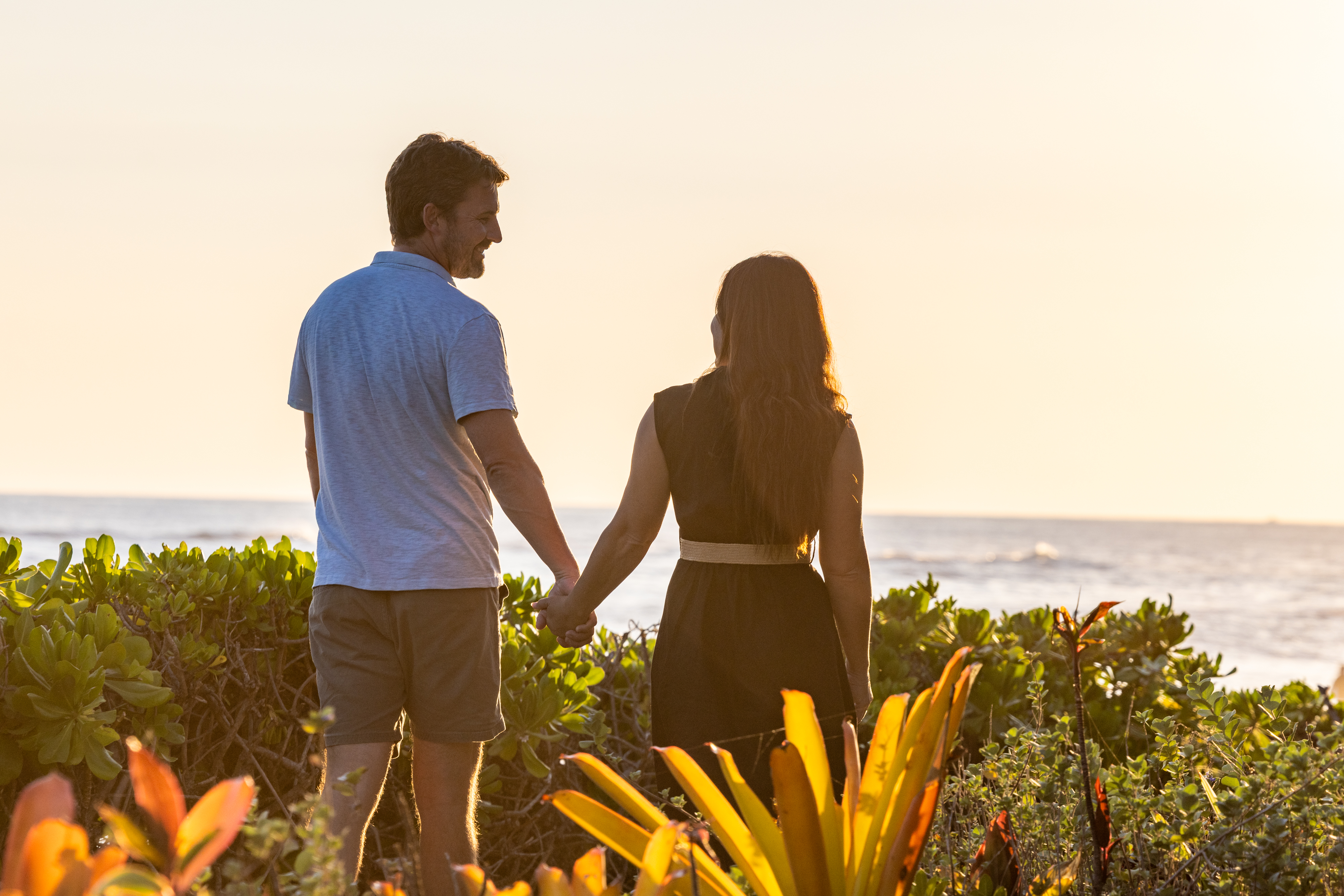 A man and woman hold hands while standing near lush plants, facing the ocean at sunset during a Manager's Meet and Greet.
