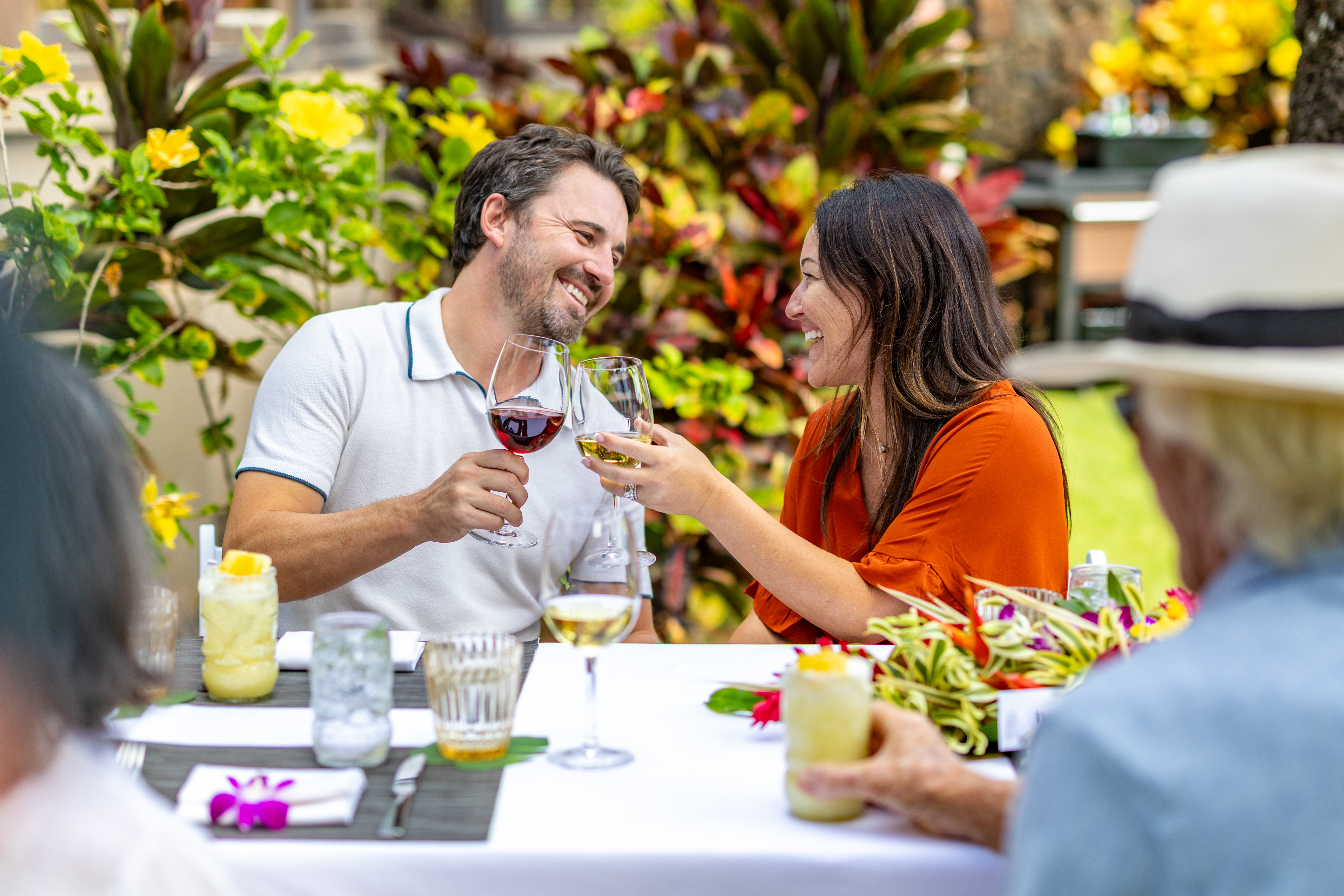 A man and woman sit at an outdoor table clinking wine glasses and smiling, surrounded by plants and other people.