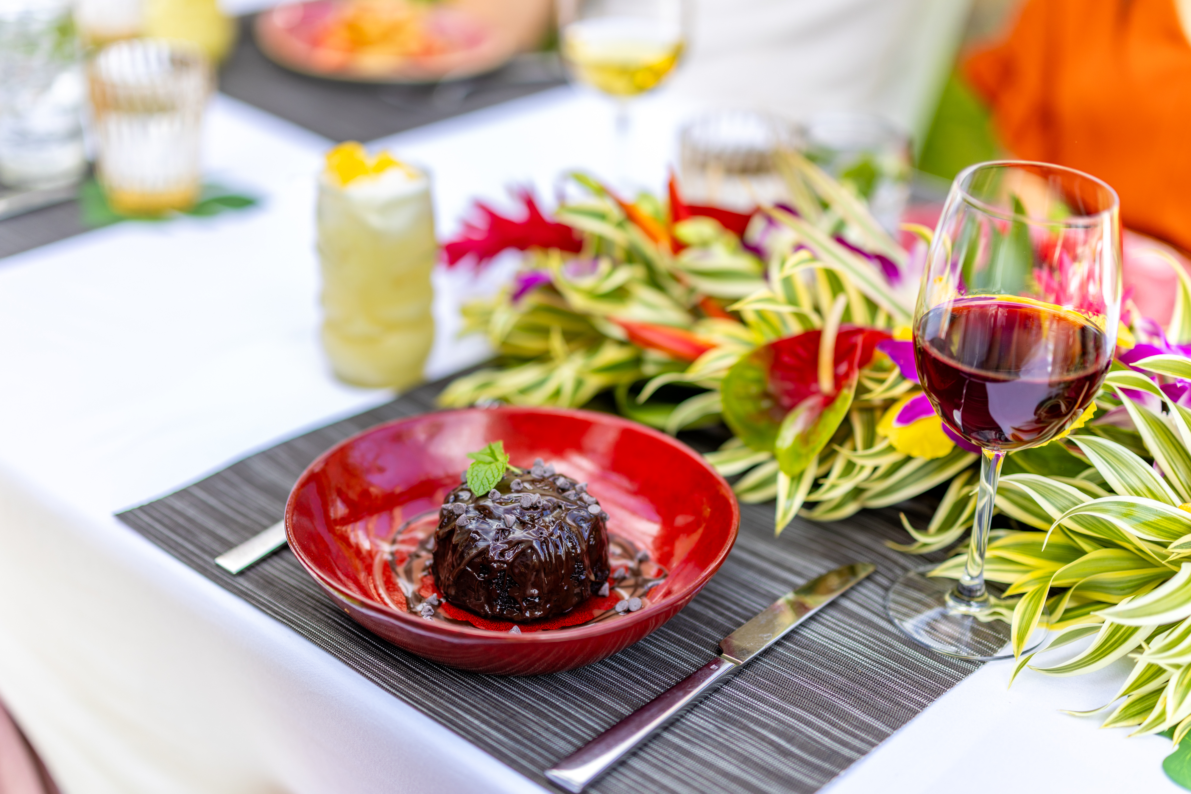 A chocolate dessert sits in a red bowl on a set table, accompanied by a knife and fork, a glass of red wine, tropical floral arrangement, and drinks in the background—capturing the festive spirit of Paina Dinners.