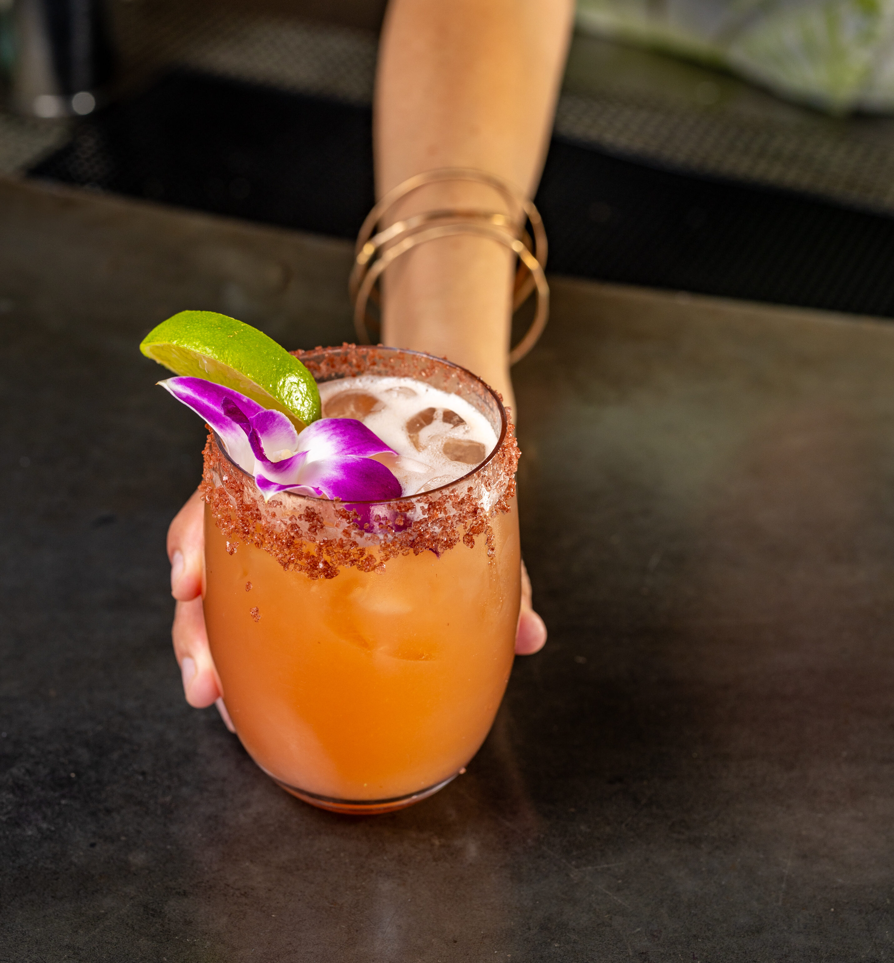 A person hands over a cocktail with a salted rim, garnished with a lime wedge and a purple orchid flower, on a dark countertop.