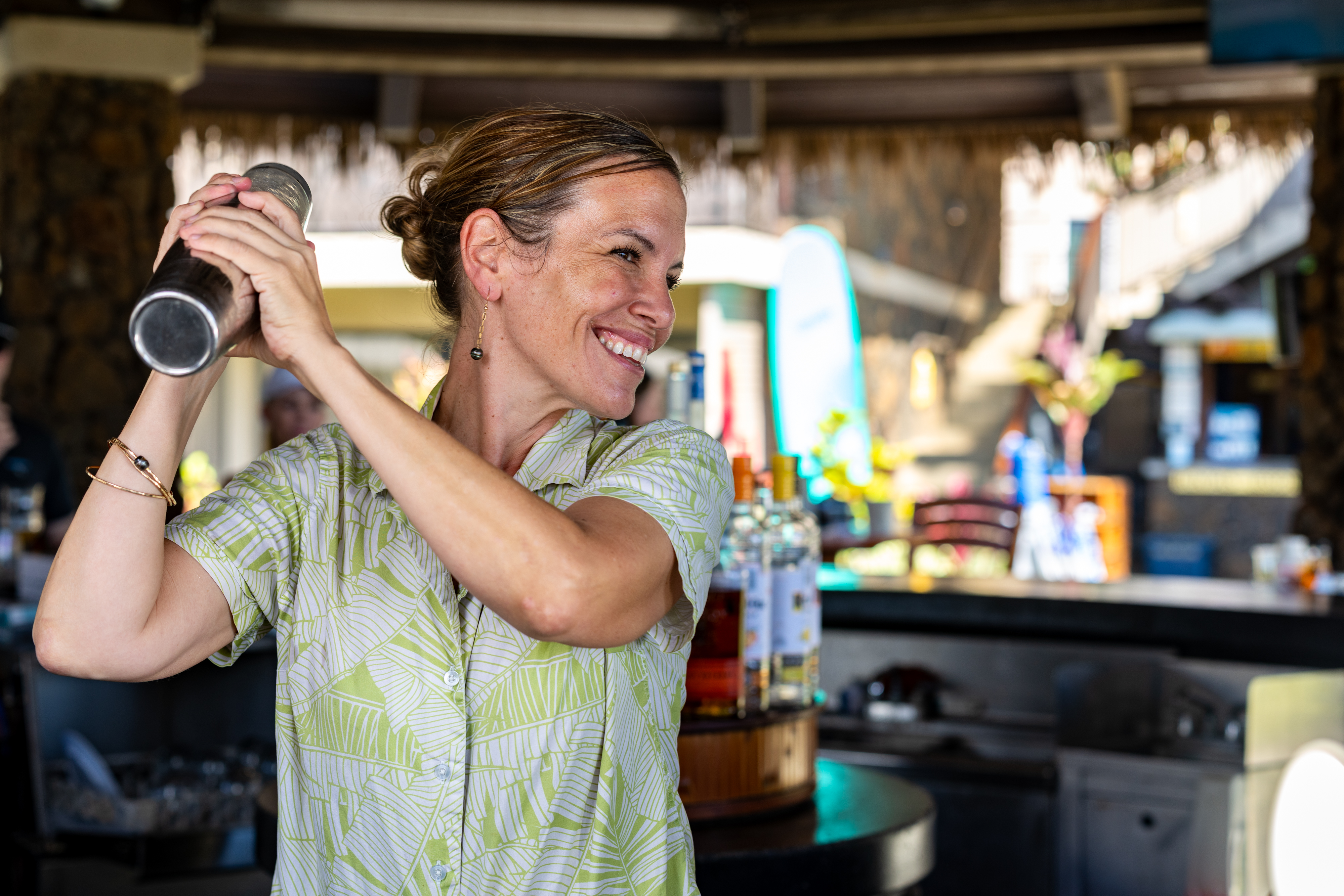 A woman smiling and shaking a cocktail shaker behind a bar, with bottles and bar equipment visible in the background.