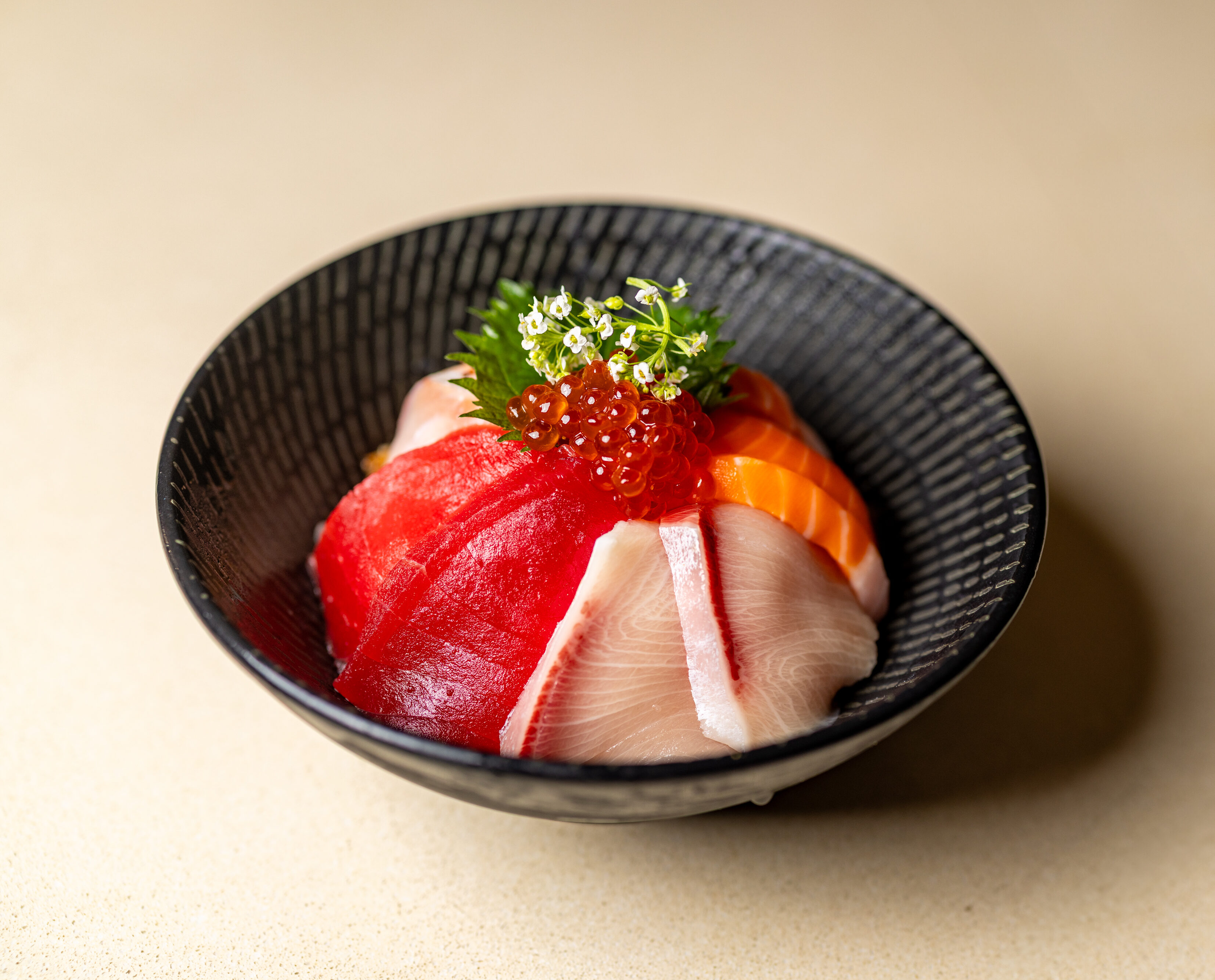 A black bowl contains assorted sashimi slices, salmon roe, and garnishes, arranged neatly on top of rice.