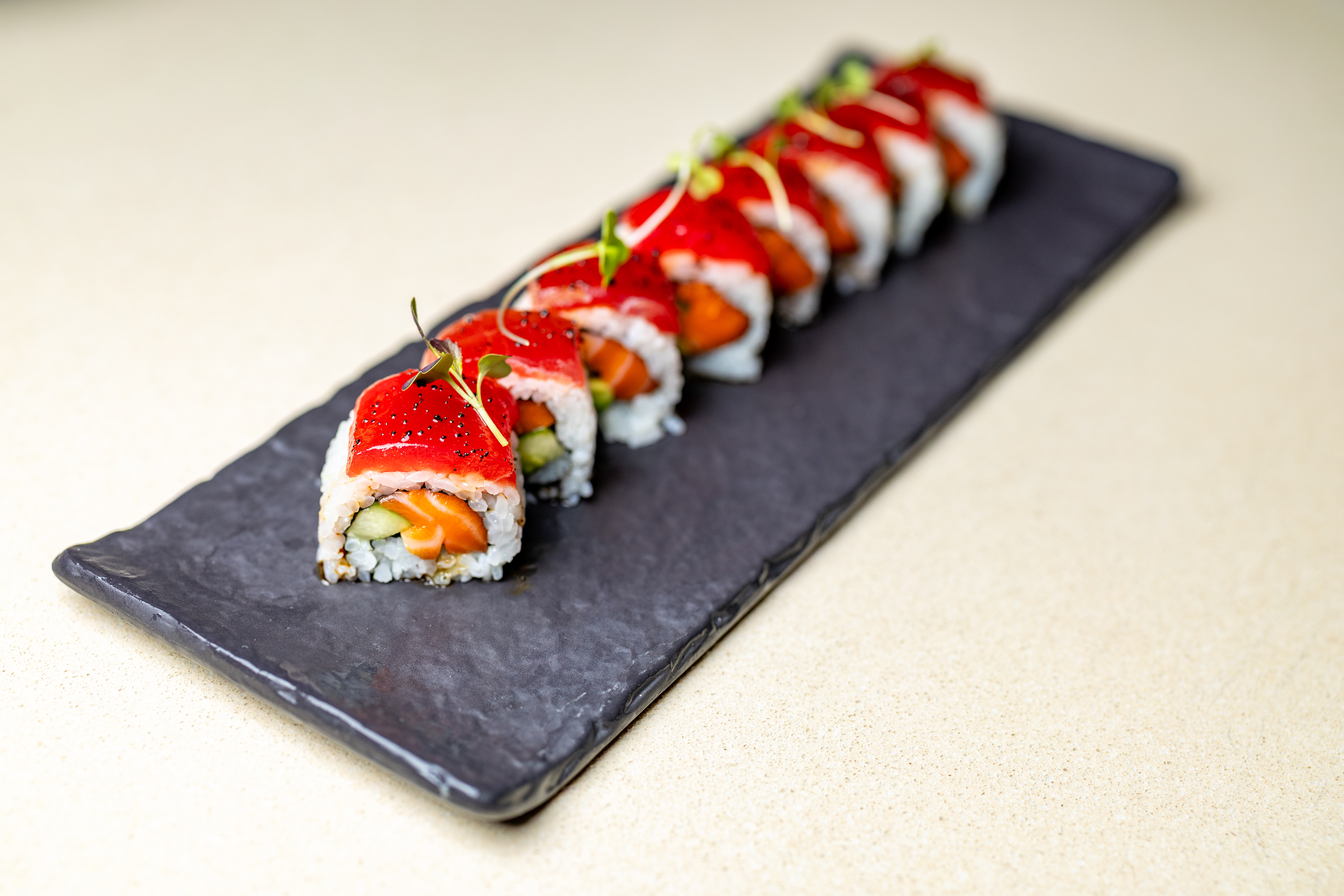 A row of sushi rolls with red topping, sliced and arranged neatly on a rectangular black plate against a light background.