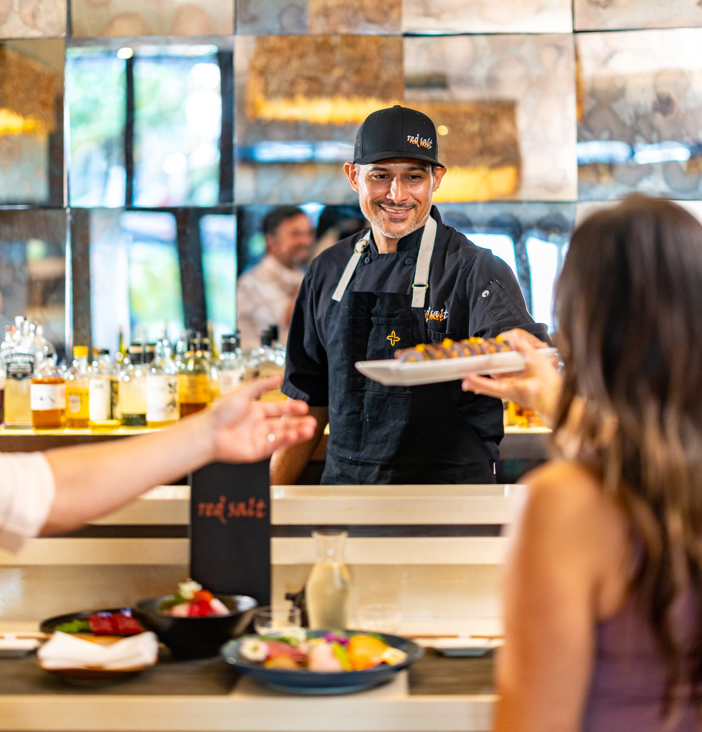 A chef in a black uniform and hat hands a plate of food to a seated guest across the counter in a modern restaurant. Other dishes and drinks are visible on the counter.