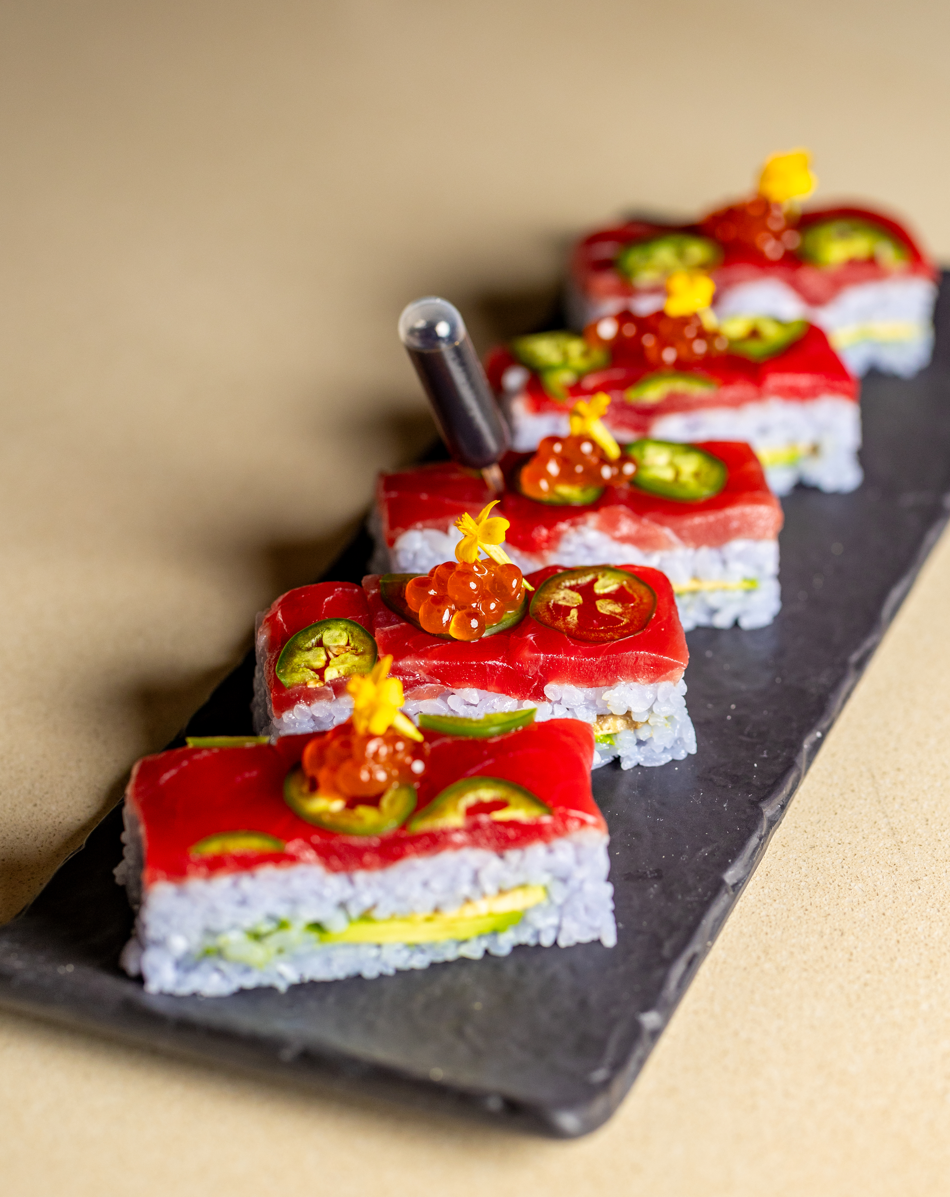 Rectangular sushi pieces layered with tuna, rice, avocado, and topped with sliced jalapeños, salmon roe, and edible flowers on a slate serving board.