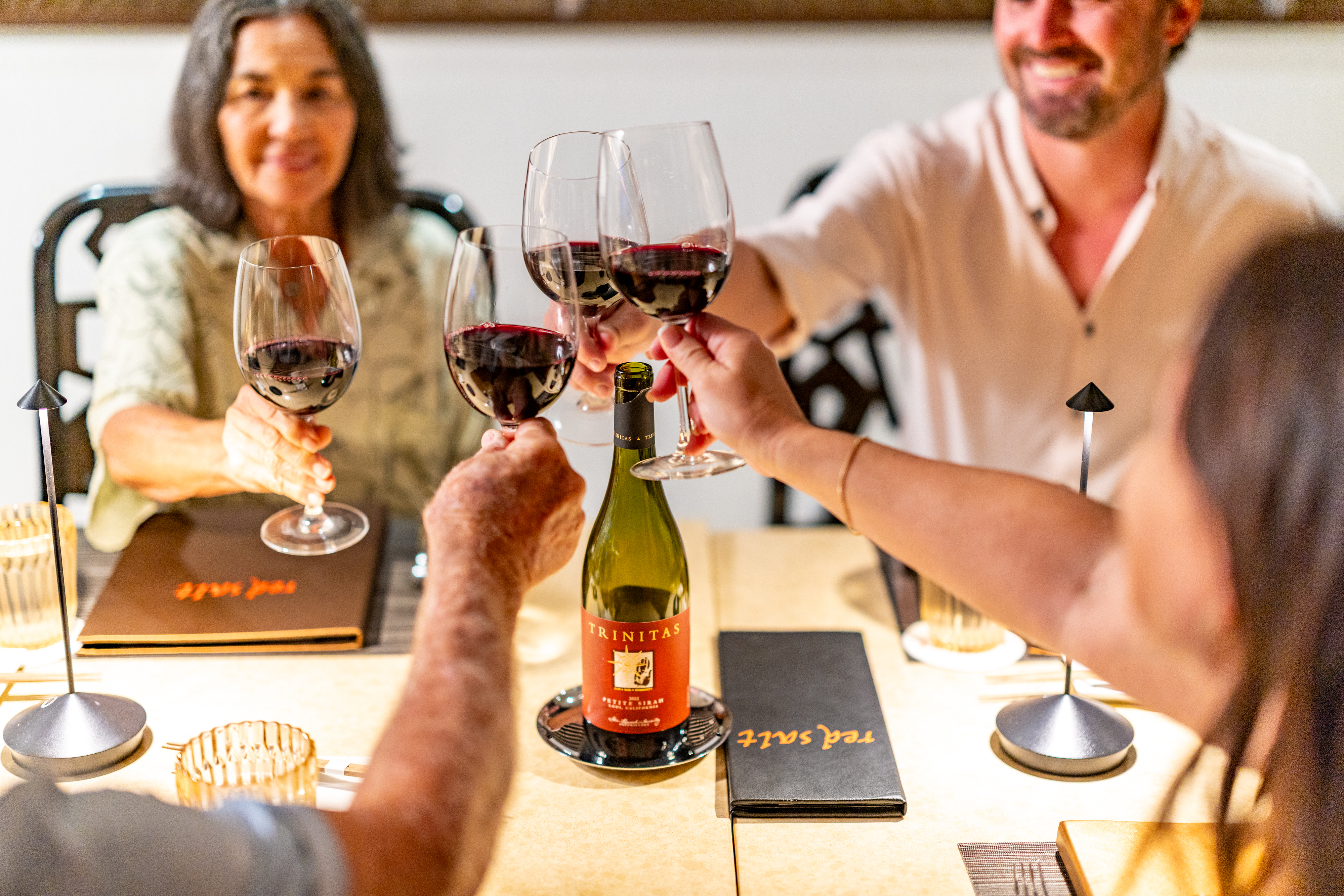 Four people clinking glasses of red wine in celebration over a table with a bottle of Trinitas wine, menus, and small lamps.