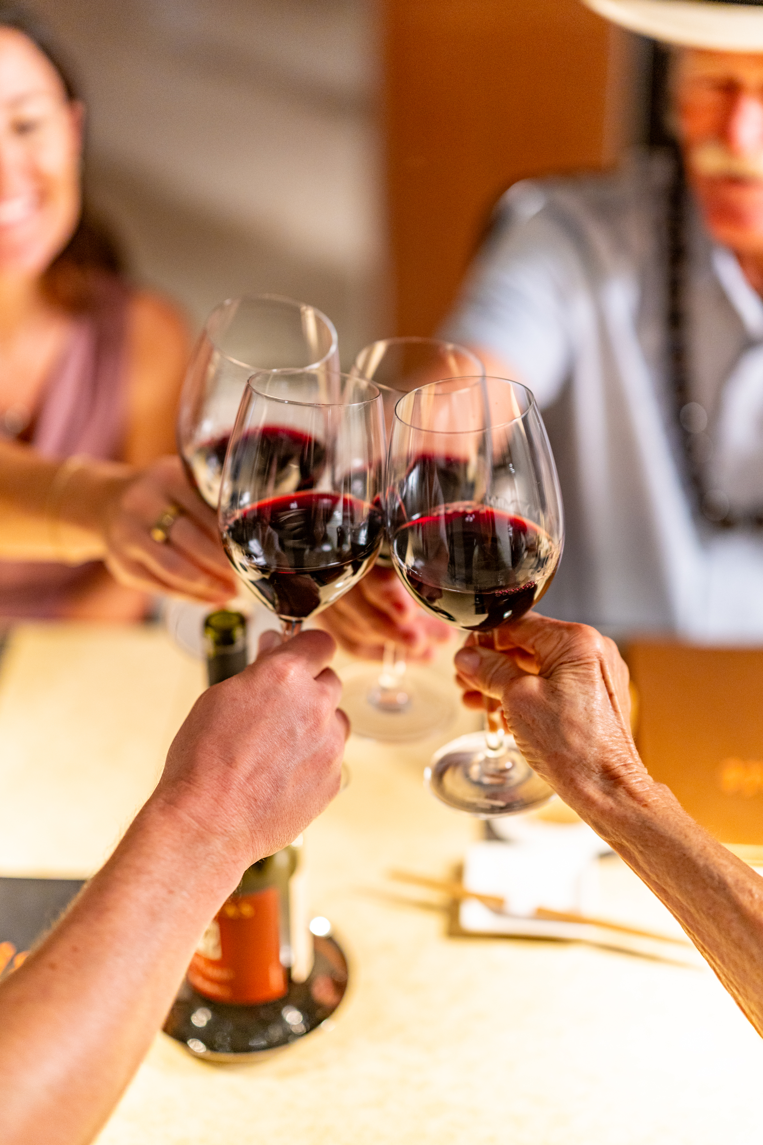 Four people clinking glasses of red wine over a table with a wine bottle and menu visible, celebrating or toasting.