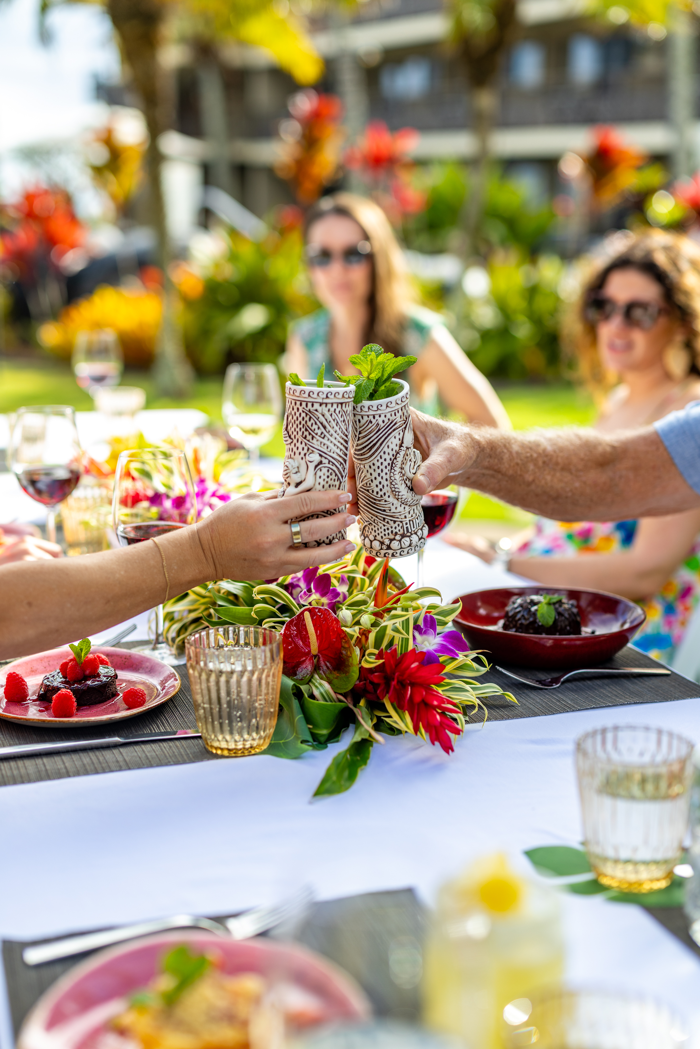 People clink decorative glasses at an outdoor table set with desserts, drinks, and flowers, with blurred individuals and greenery in the background.