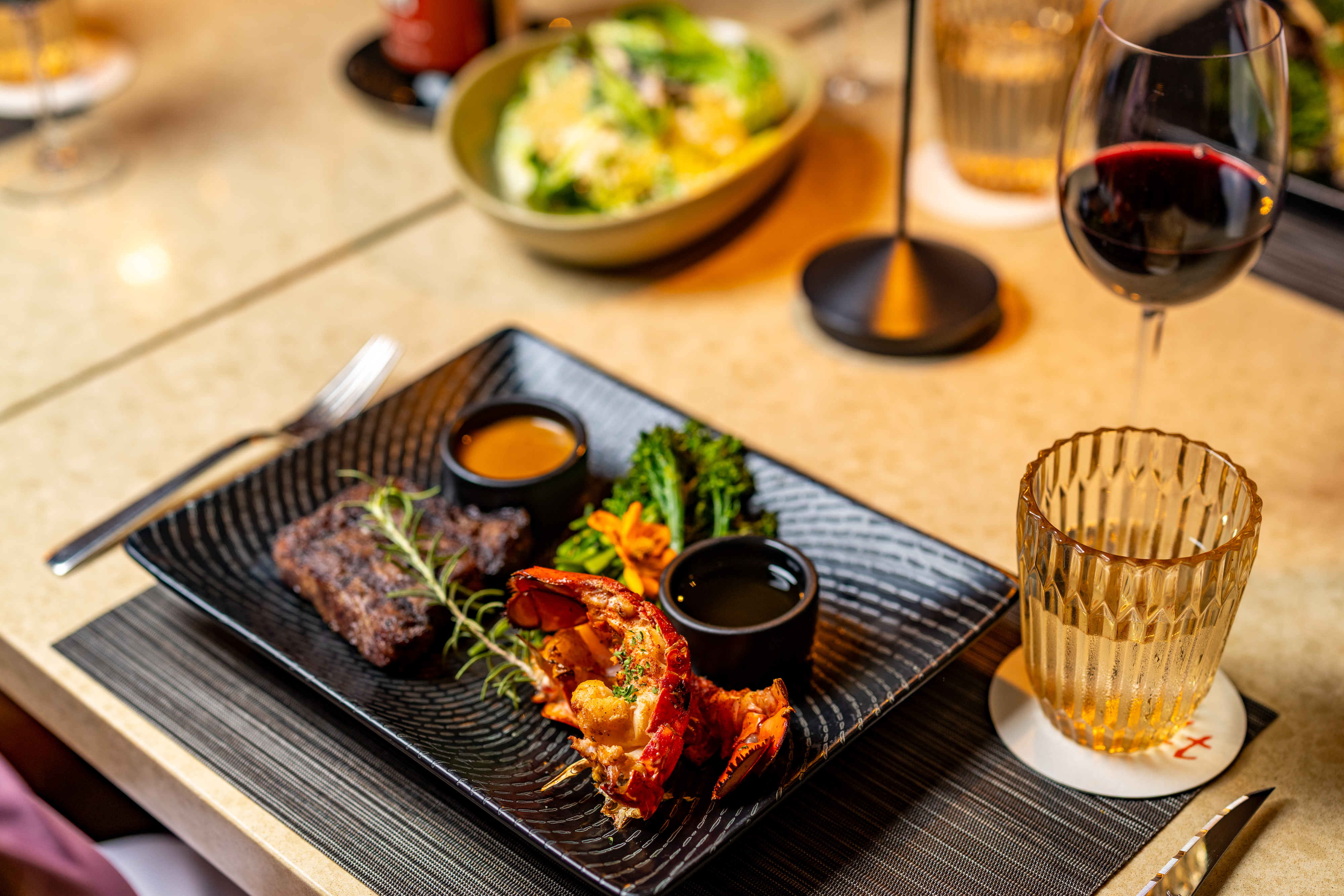 A restaurant table setting with a black plate featuring steak, lobster tail, broccolini, sauces, a glass of red wine, and a glass of water. A bowl of salad is in the background.