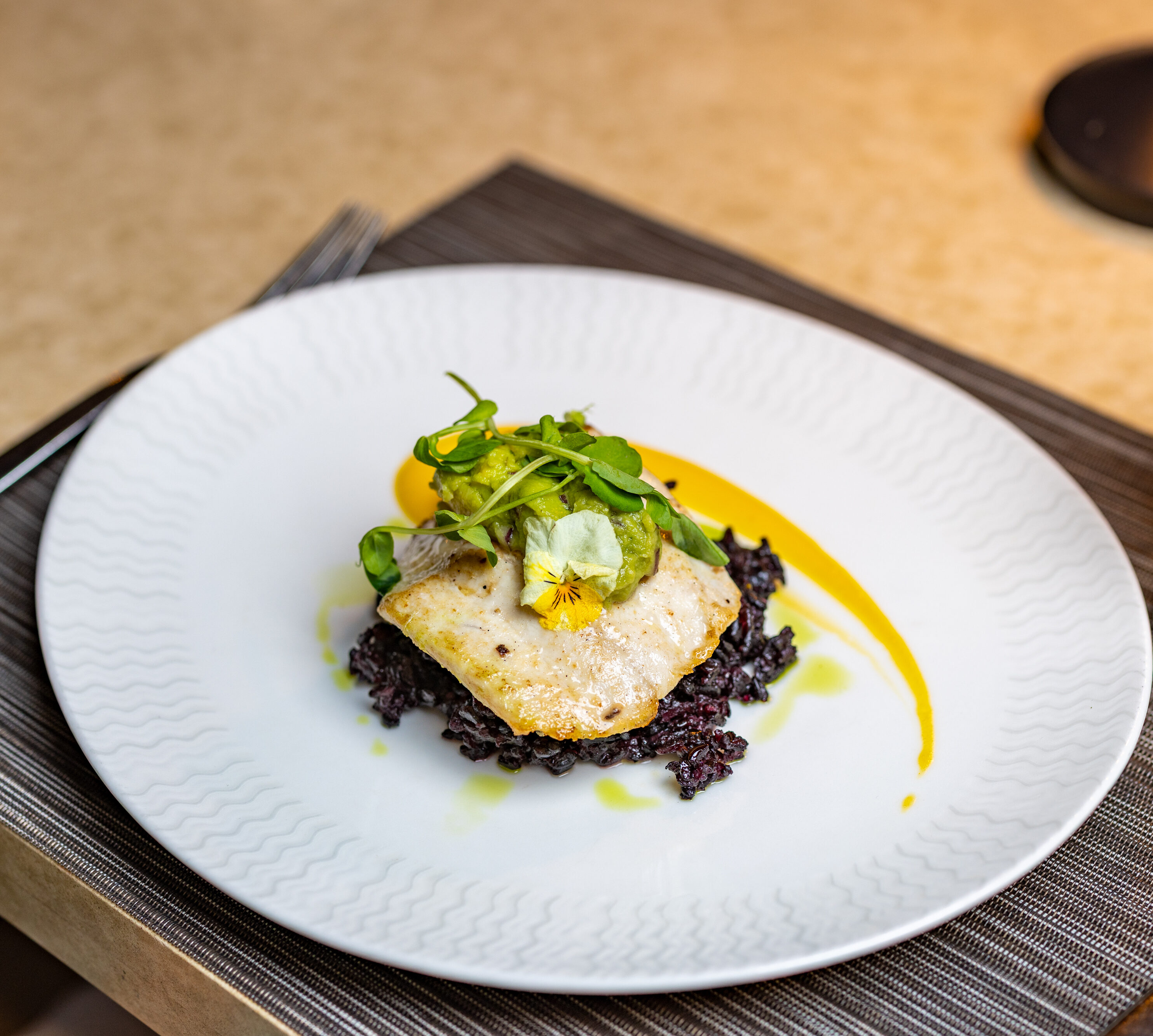 A plated dish featuring a fillet of white fish on a bed of black rice, garnished with greens and a yellow sauce, set on a table with a glass of water and a placemat.