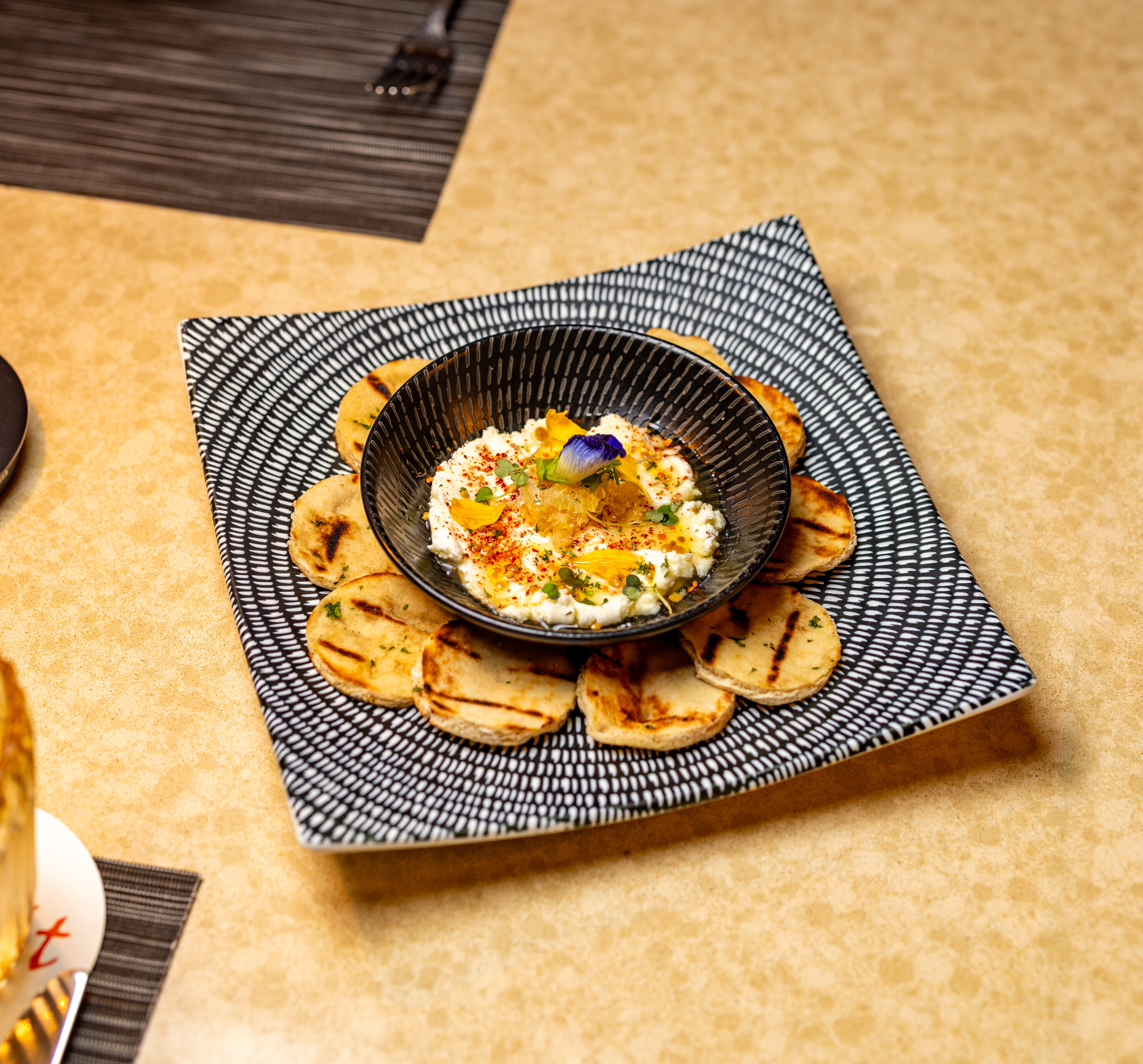 A bowl of dip garnished with herbs and edible flowers, surrounded by grilled pita slices, is served on a patterned plate on a beige table.
